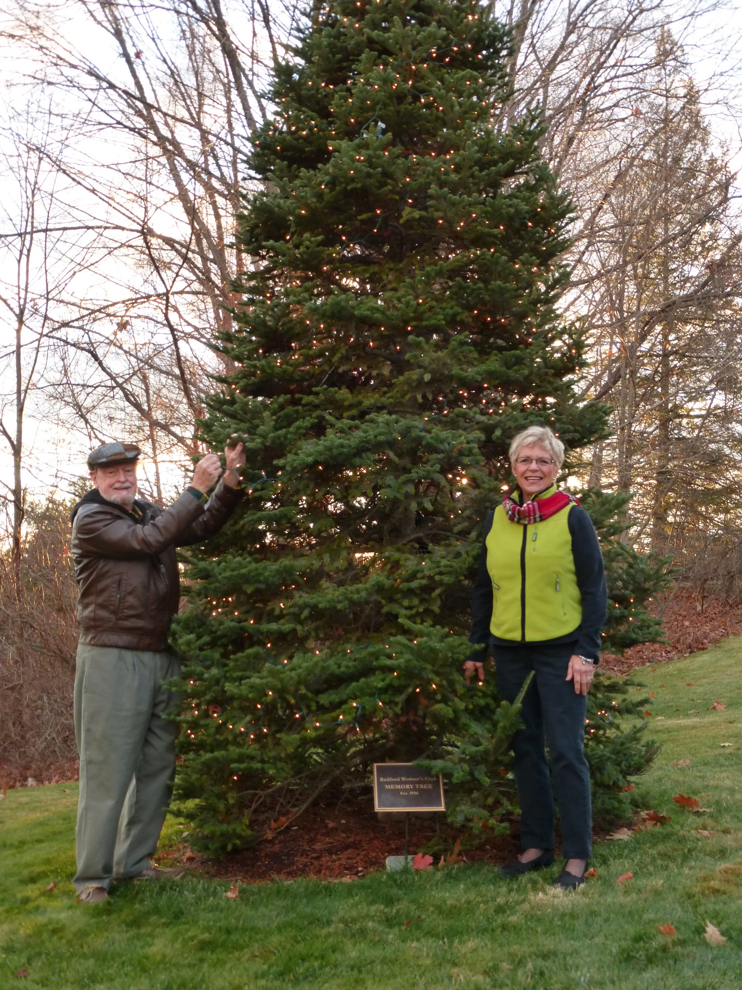 A man and woman stand next to a tall, decorated Christmas tree outdoors during twilight. The man is wearing a leather jacket, glasses, and a flat cap, and is holding a string of lights. The woman is wearing a yellow vest over a dark long-sleeve shirt, glasses, and a red scarf, and they are smiling at the camera.