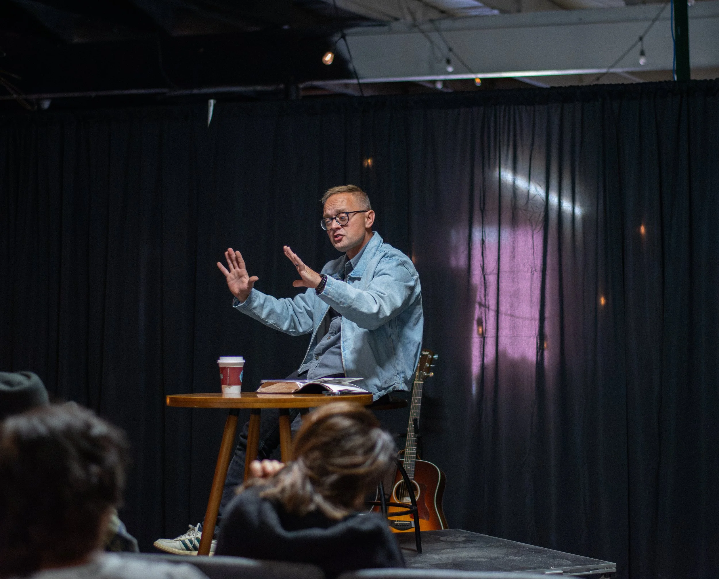 A man with glasses and a denim jacket speaking on stage during a performance or talk, with a guitar and a cup on a wooden table in front of him, set against dark curtains and lighting during an indoor event.