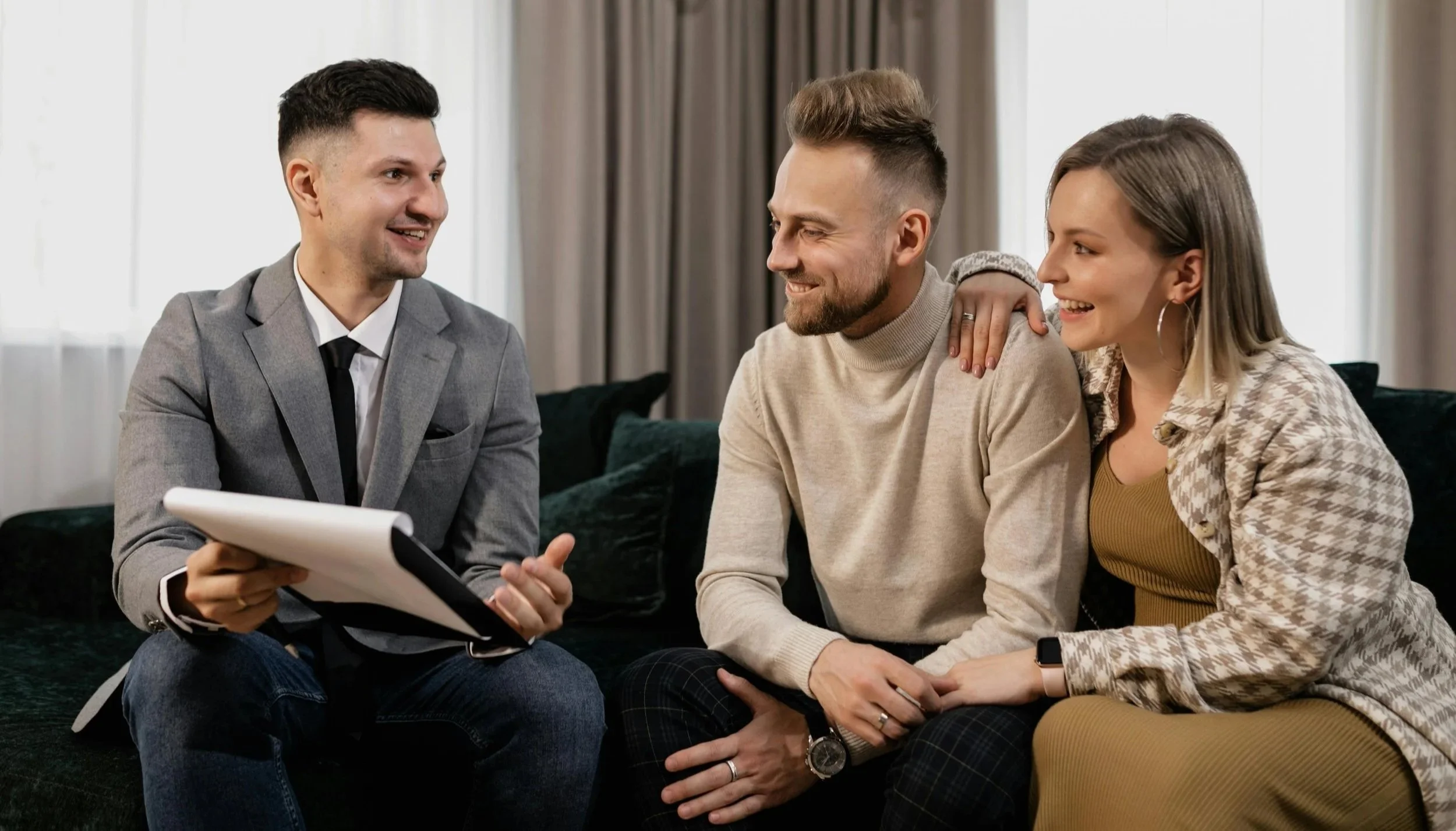 A man in a gray suit and tie consulting with a couple on a couch. The couple includes a man in a beige sweater and a woman in a patterned shirt, all smiling and engaging in conversation.