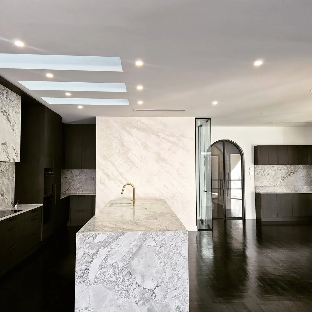 Modern kitchen with black cabinets, marble island, gold faucet, skylights, wooden flooring, and a glass door with an arched window.