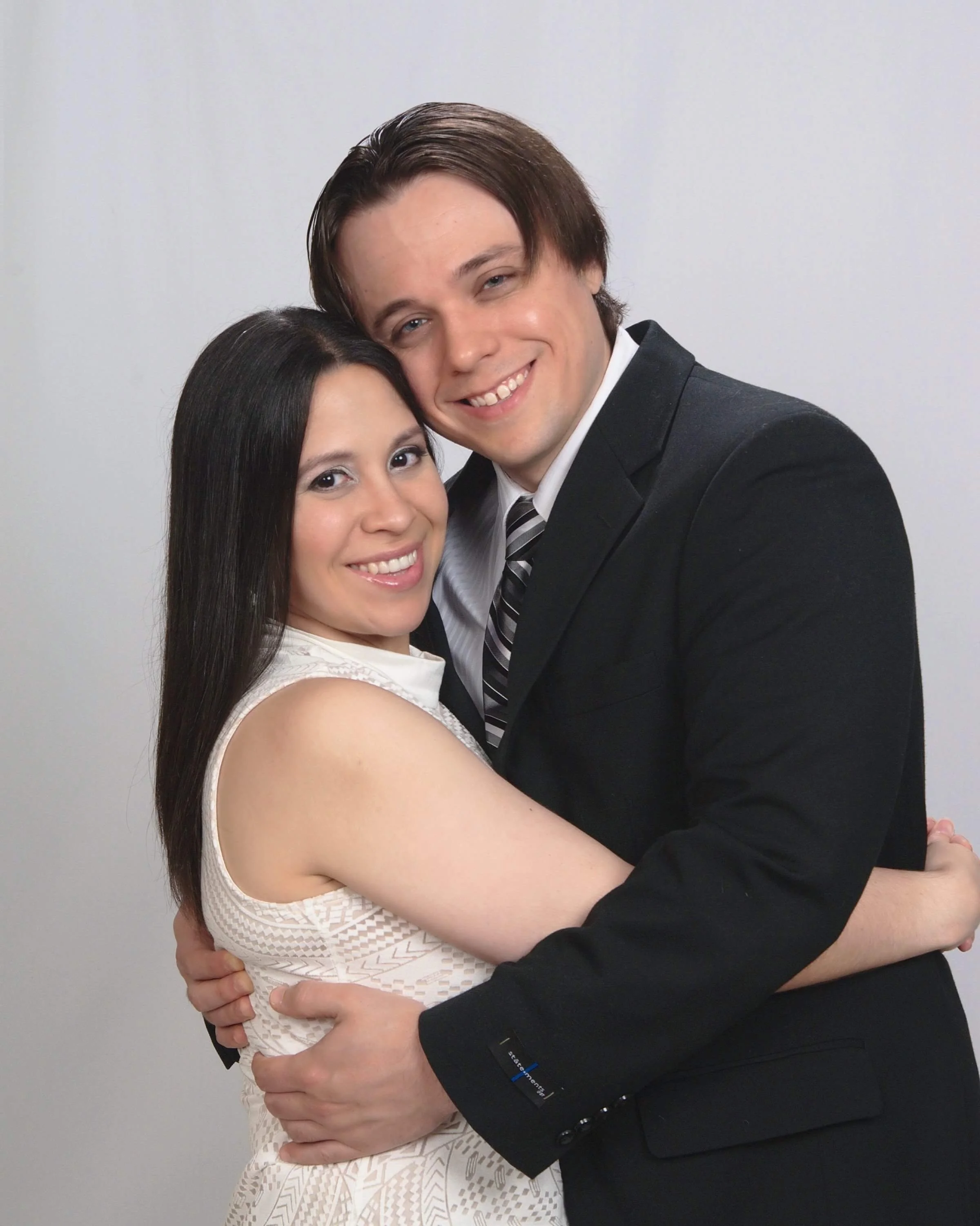 A smiling young woman with long dark hair wearing a sleeveless white dress, embracing a young man with medium-length dark hair wearing a black suit, white shirt, and striped tie, both standing against a plain light-colored background.