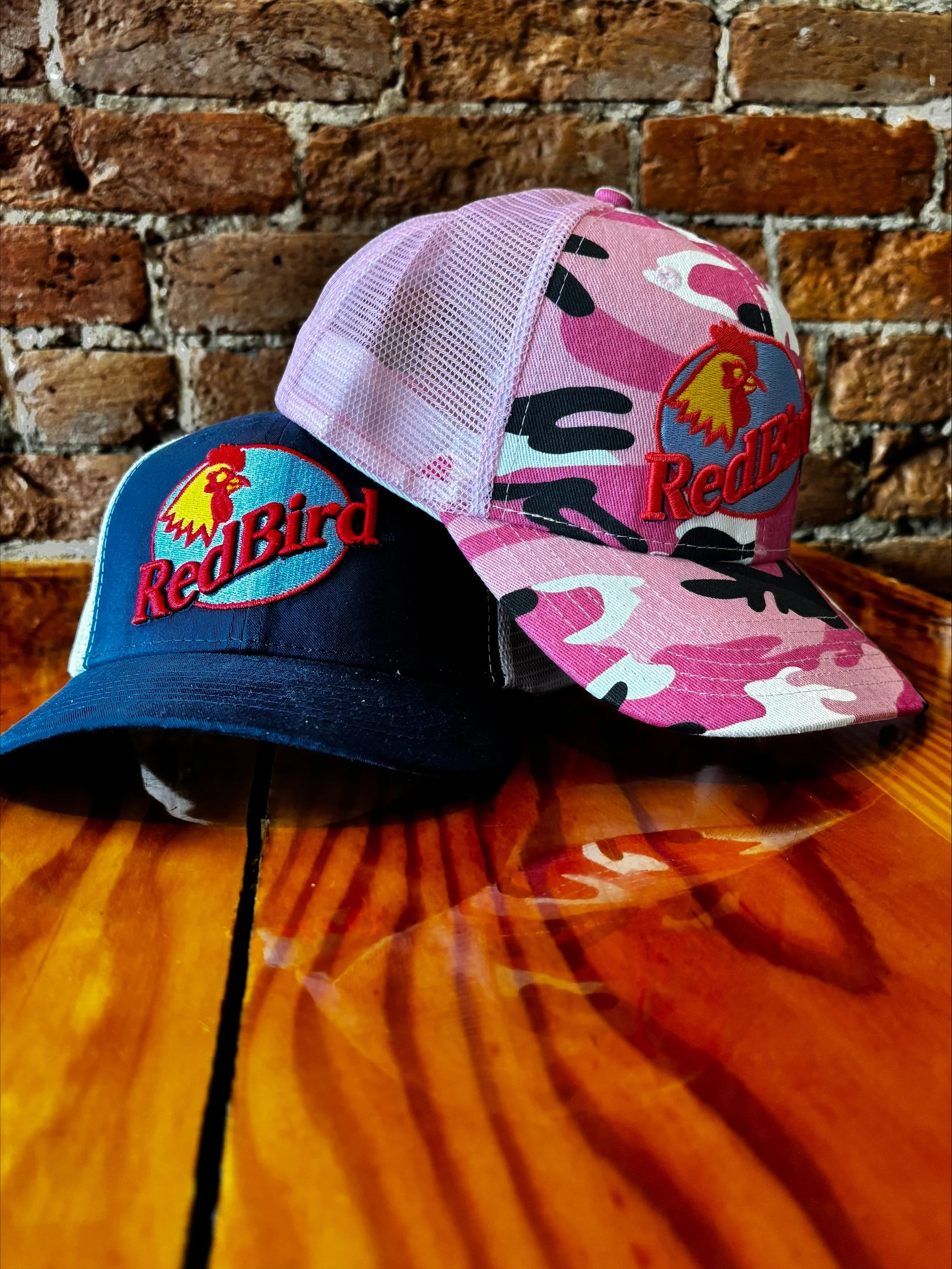 Two baseball caps on a wooden table with a brick wall in the background. One cap has a pink camouflage pattern with a 'Red Bird' logo, and the other is dark blue with a matching logo.