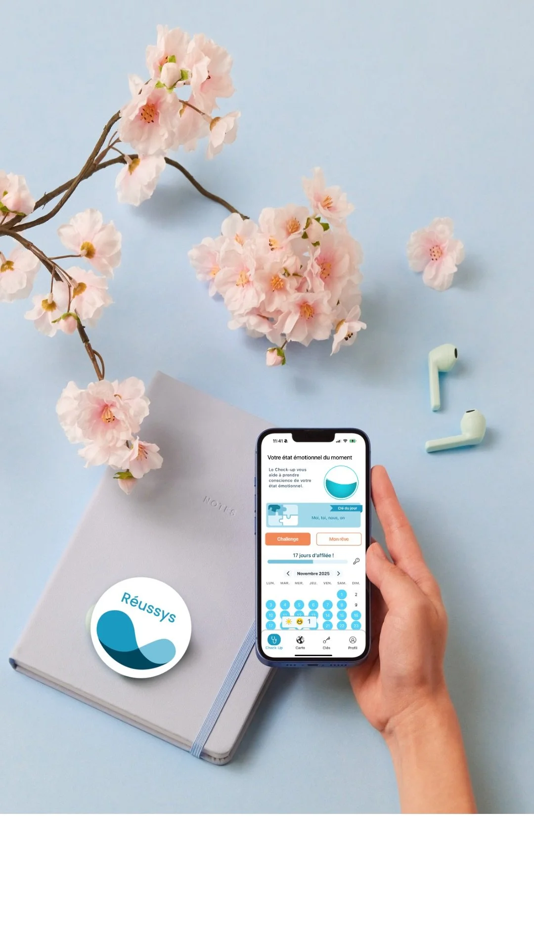A person holding a smartphone with an mental health tracking app open, surrounded by pink cherry blossoms, wireless earbuds, and a gray notebook with a sticker that says 'Réussys' on a light blue background.