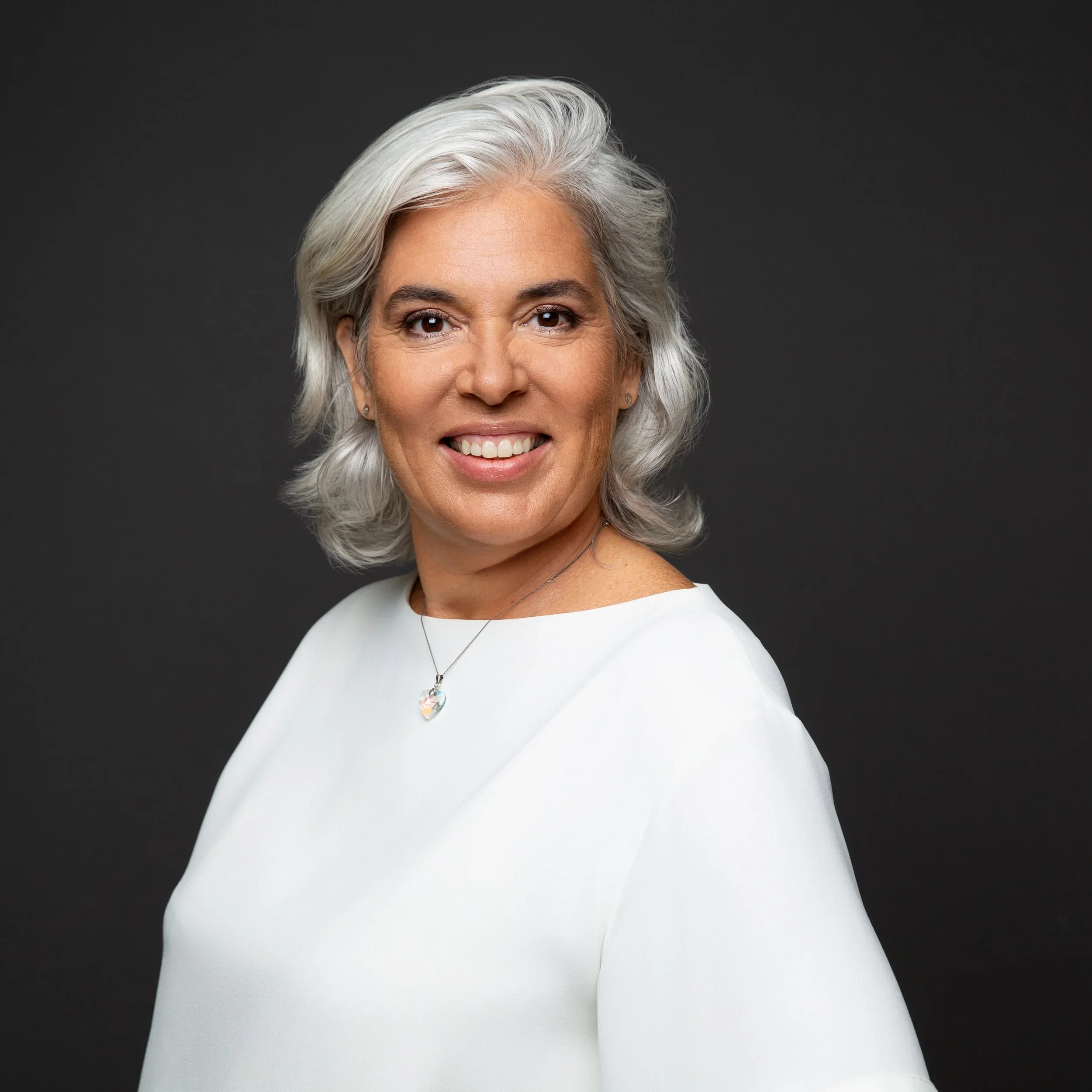 A smiling woman with gray hair, wearing a white top and a necklace with a heart-shaped pendant, against a dark background.