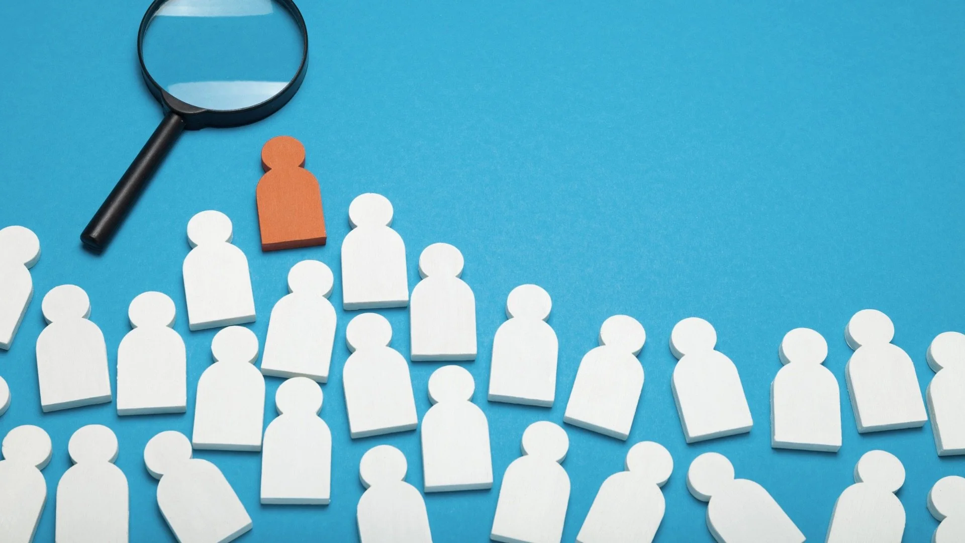 A magnifying glass, a brown human-shaped figure, and several white human-shaped figures arranged on a blue surface, suggesting a concept of searching or analyzing a specific individual among a group.