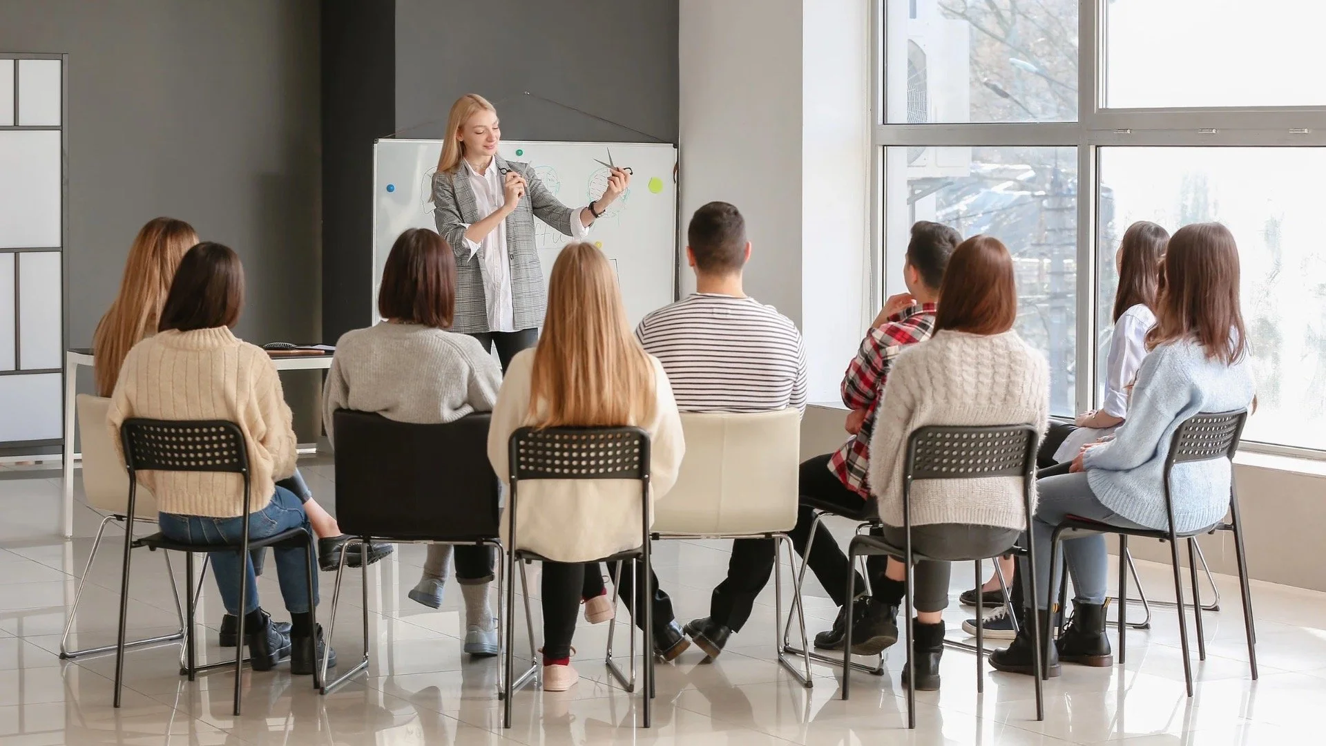 A woman teaching a class to a group of young adults seated in a semi-circle in a bright classroom with large windows.