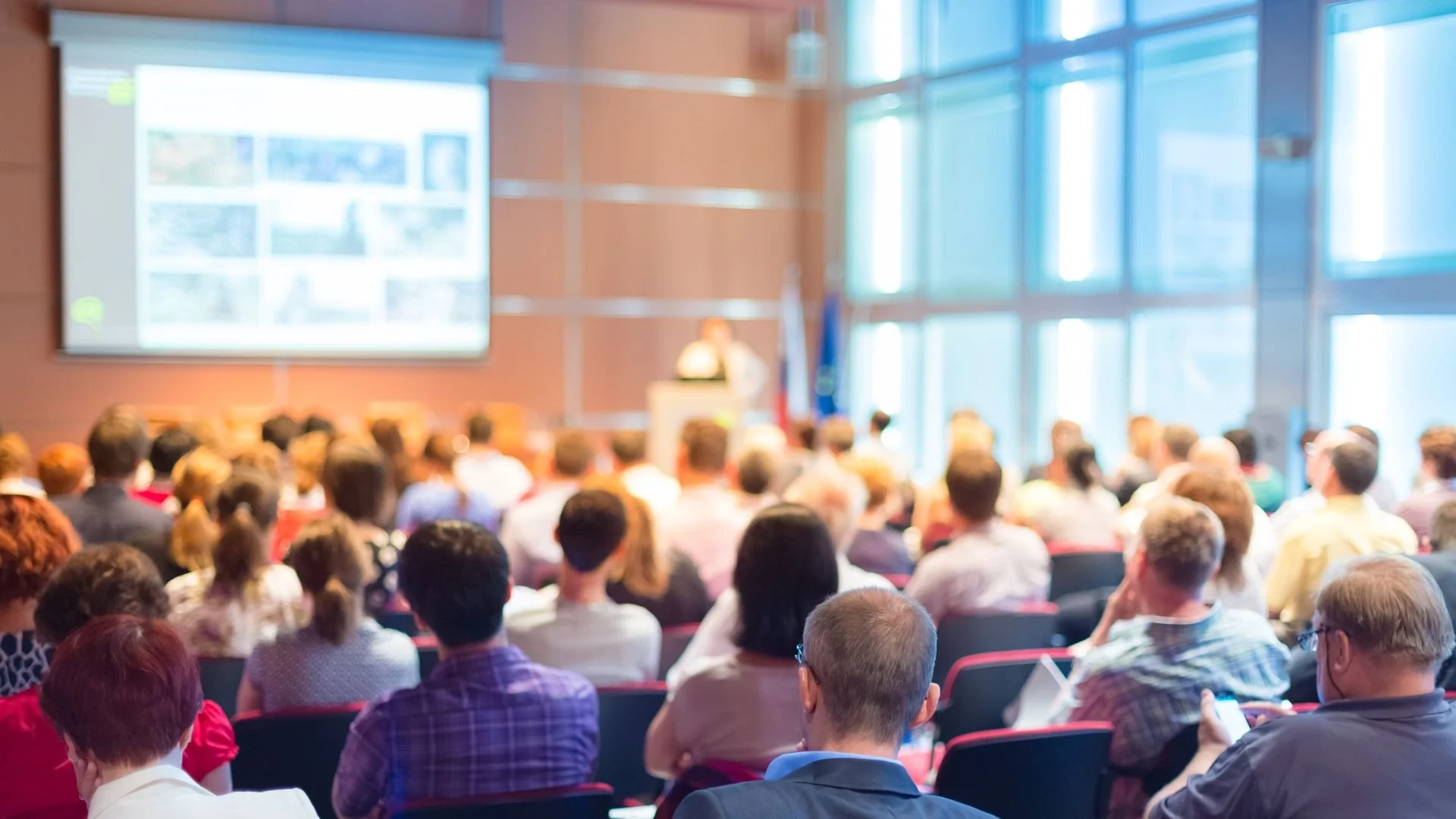 Audience attending a conference or seminar in a bright, modern room with large windows, facing a speaker at a podium and a presentation screen.