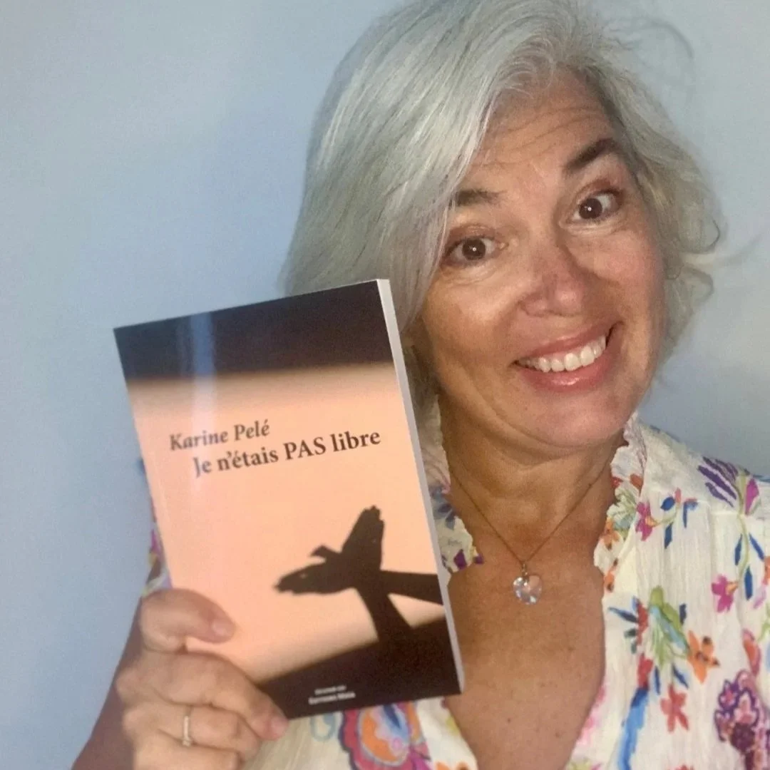 A smiling woman with gray hair holding a book titled "Je n'étais PAS libre" by Karine Pelé, standing against a plain light-colored wall.