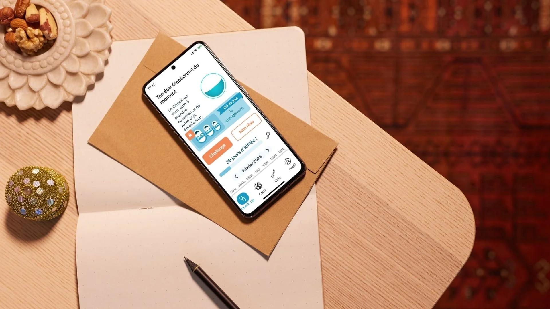 A smartphone on top of a closed brown notebook and a white notebook, placed on a wooden desk. The screen displays a French language app with emotional health tracking. To the left, there is a notebook, a decorative golden ball, and to the top, part of a white decorative plate with nuts. A black pen is lying on the white notebook.