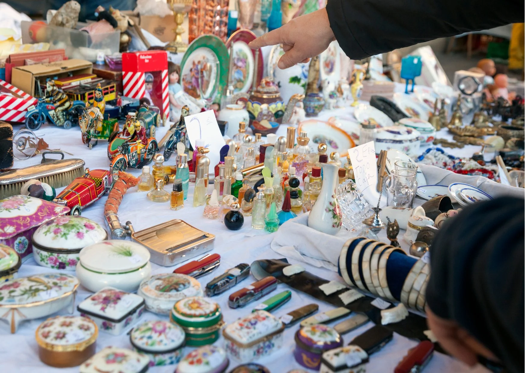 A market stall with vintage items including decorative boxes, perfume bottles, jewelry, and miniature carousel figurines; a person's hand is pointing toward the items.