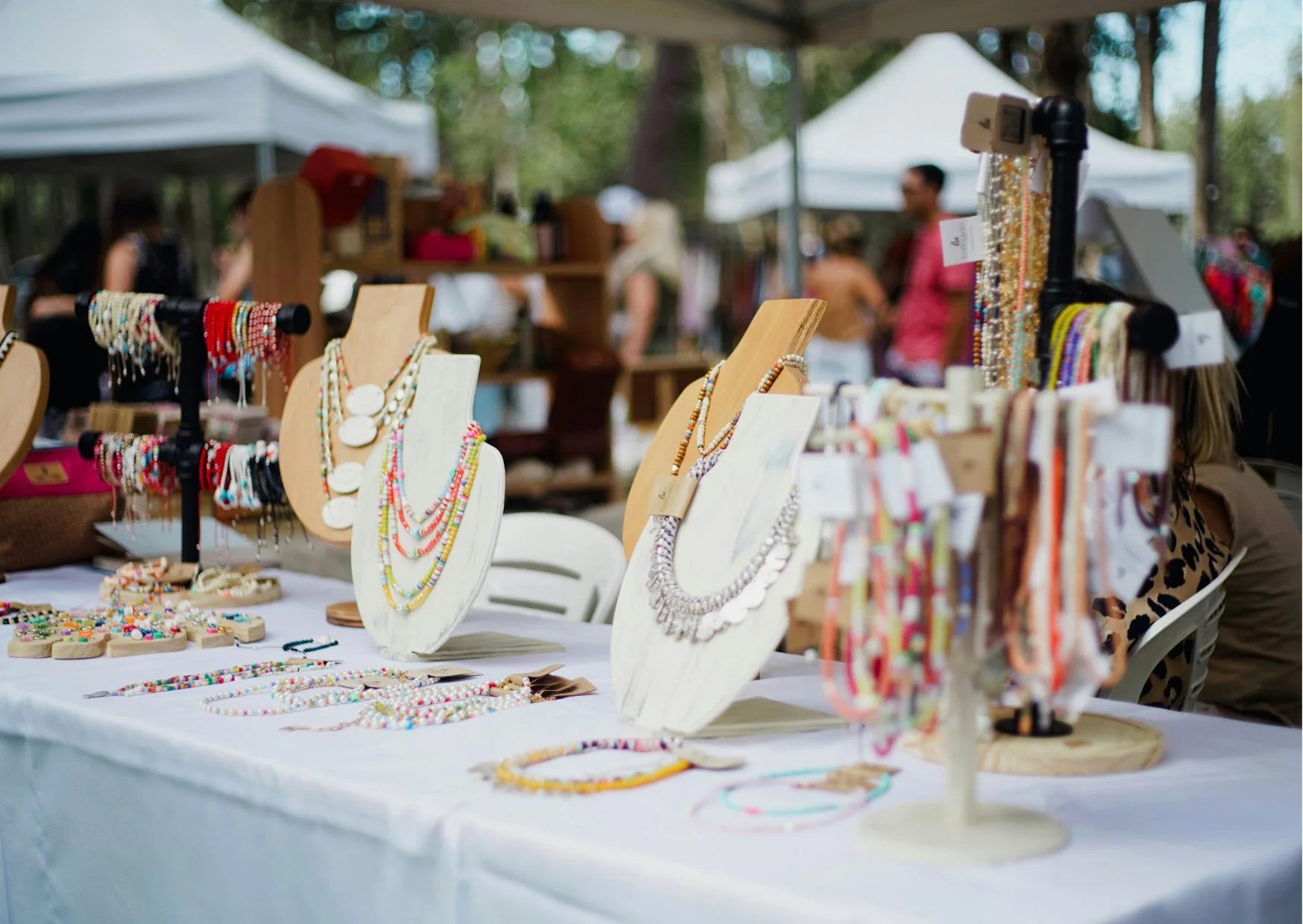 Jewelry displayed on a table at an outdoor market, with necklaces and bracelets on wooden and black stands, under white tents with people browsing in the background.