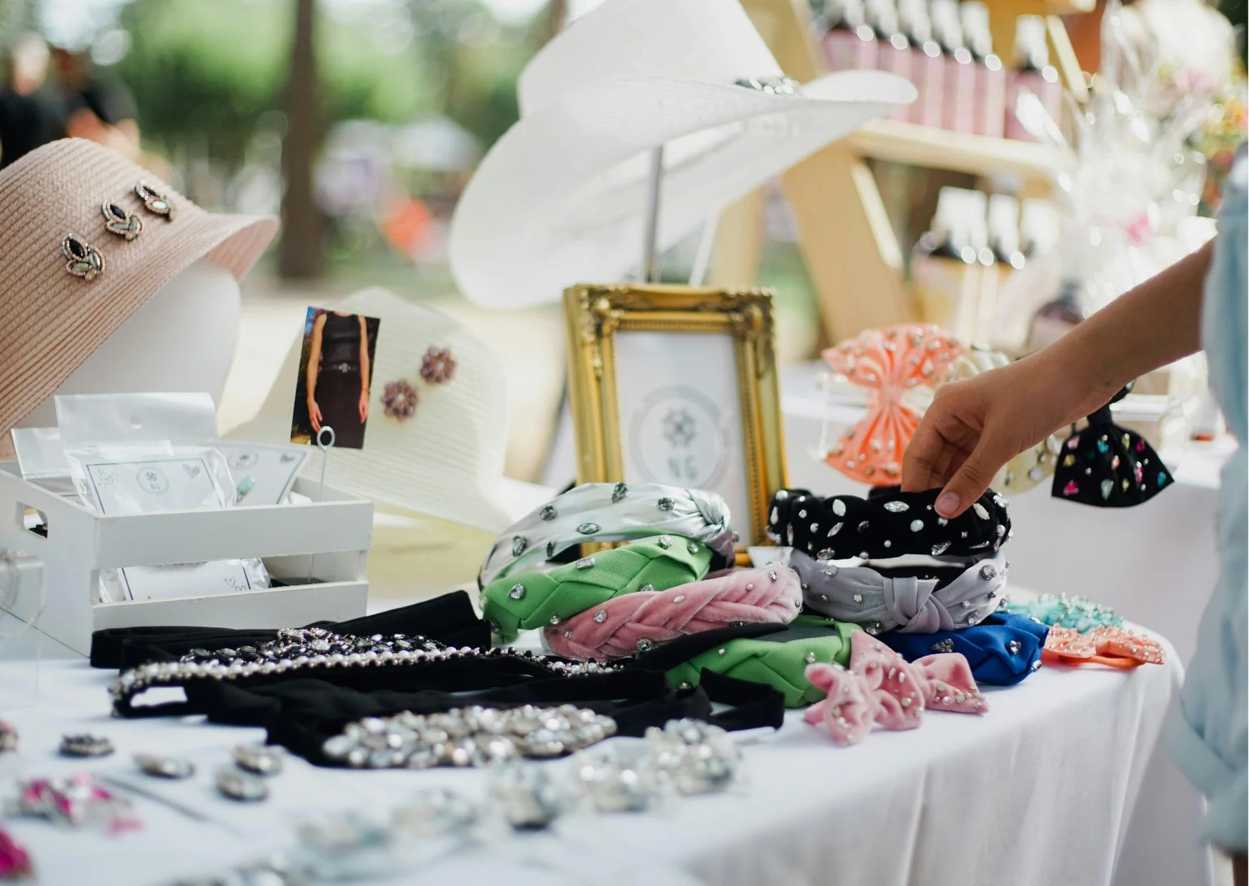 A table at an outdoor market displays various accessories such as headbands with jewels, hats with pins, earrings, and bracelets in different colors and styles. A person's hand is reaching for a black headband with white dots.