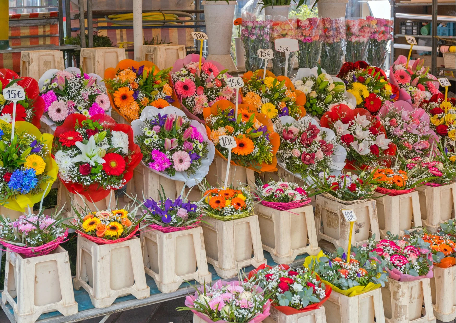 Colorful flower bouquets displayed at a market stall, with each bouquet wrapped in paper or plastic and labeled with prices in euros. The arrangement includes various types of flowers such as daisies, roses, lilies, and tulips in vibrant colors like pink, red, yellow, purple, and white.