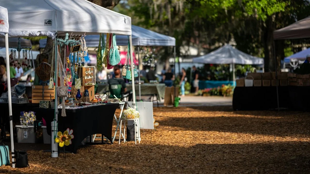 Outdoor market with various vendor tents selling handmade crafts, jewelry, and accessories. People shopping and browsing on a sunny day.