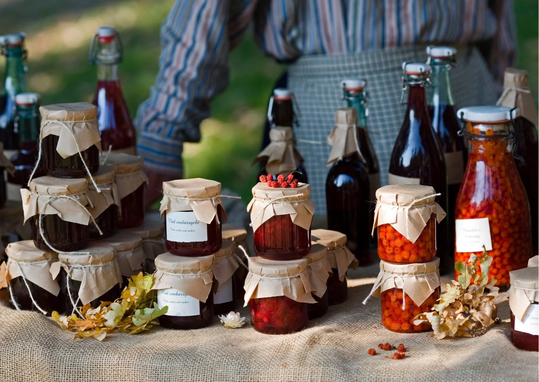 Assorted jars of homemade fruit preserves and jams on a rustic table at an outdoor market, with a person in the background wearing a striped shirt and apron.