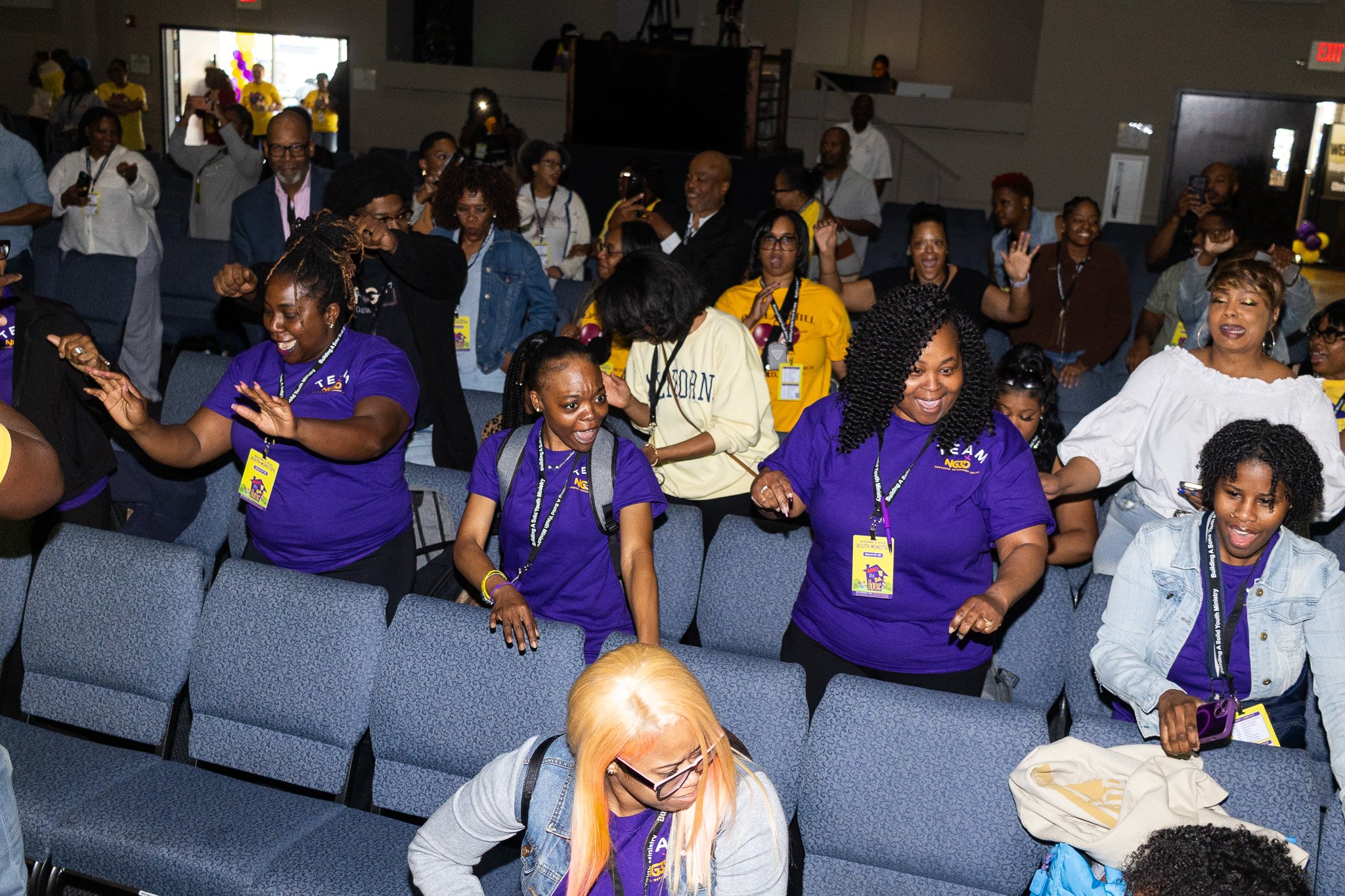 A group of diverse people attending an indoor event, some raising their hands, taking photos, or dancing. Many are wearing matching purple t-shirts and name tags.