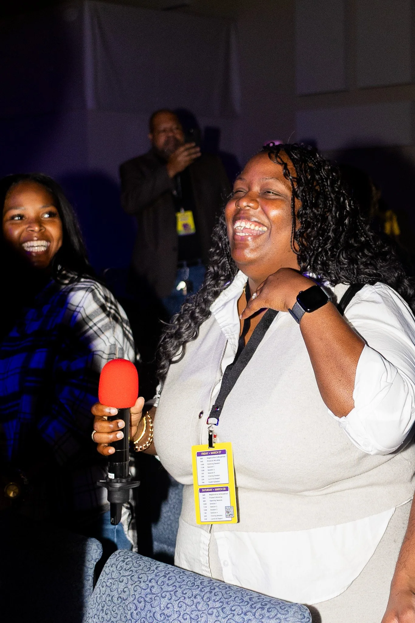 A woman with curly hair laughing and wearing a white shirt, holding a red microphone, standing in a dark indoor setting with other people in the background. Another woman is partially visible on the left, and a man is in the background taking a photo