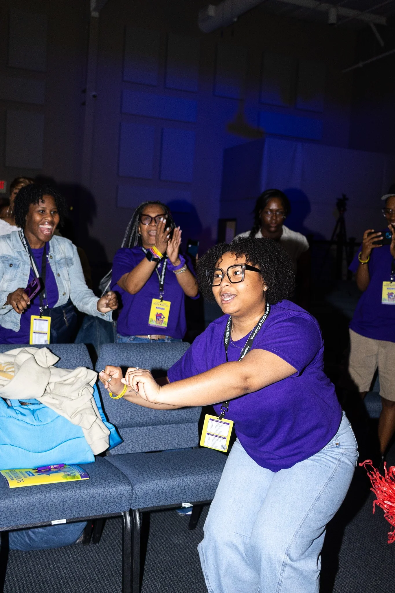 A group of people wearing purple shirts and name tags, some smiling and some taking photos, gathered around a table in a conference room with dark blue walls.