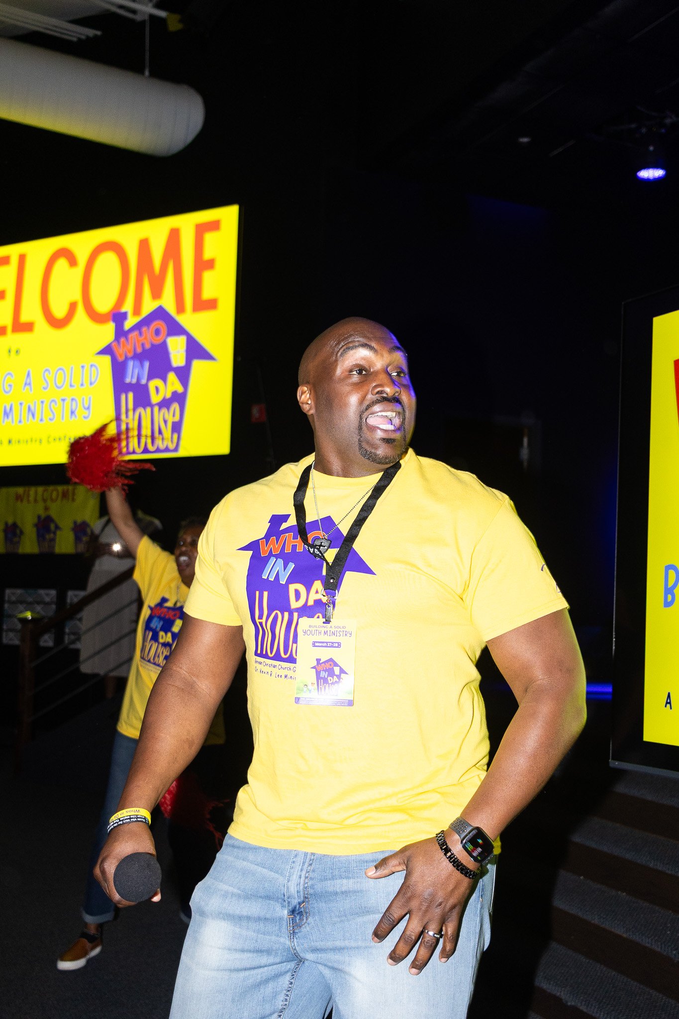 A man wearing a yellow t-shirt with a logo and a conference badge standing in a dark room. There are yellow signs with blue and purple text that say 'Welcome' and 'Who in Da House?' in the background.