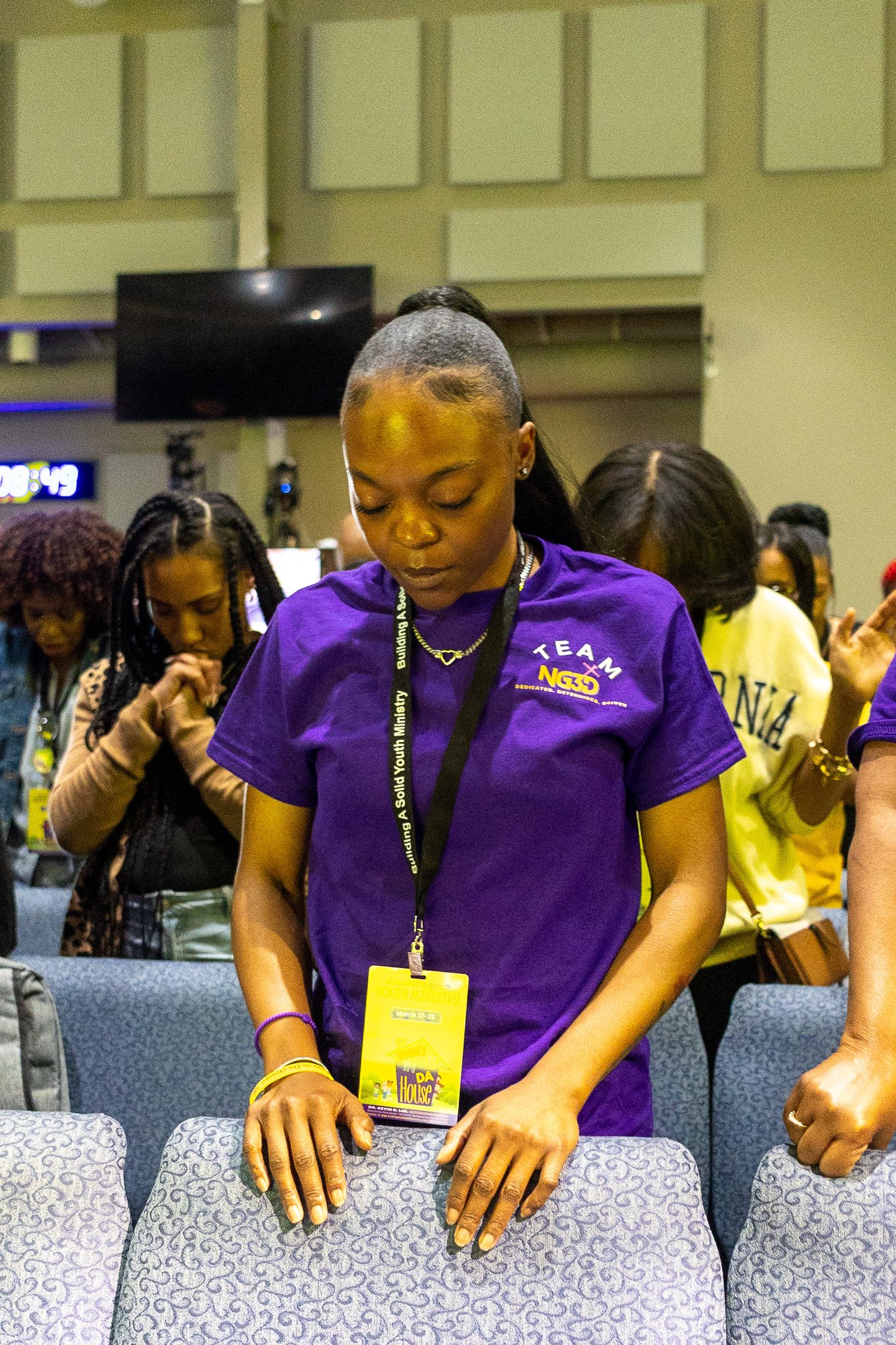 A woman with braided hair and a short haircut, wearing a purple T-shirt and a yellow lanyard, is holding her hands over a row of padded chairs with her eyes closed. Other people are visible in the background in what appears to be a conference or even