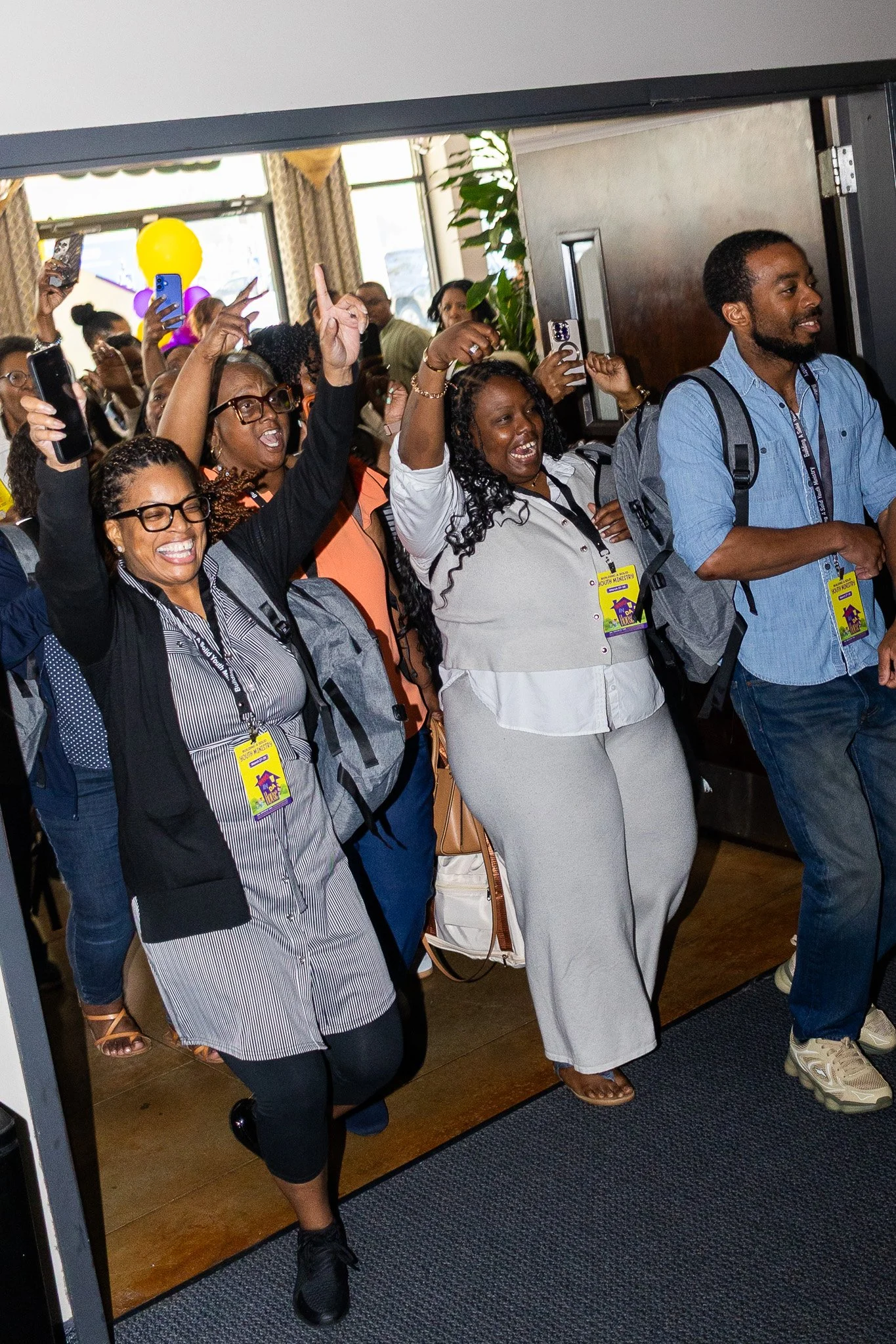 People excitedly celebrating at an indoor event, some taking photos, wearing name tags, and raising their hands in the air.