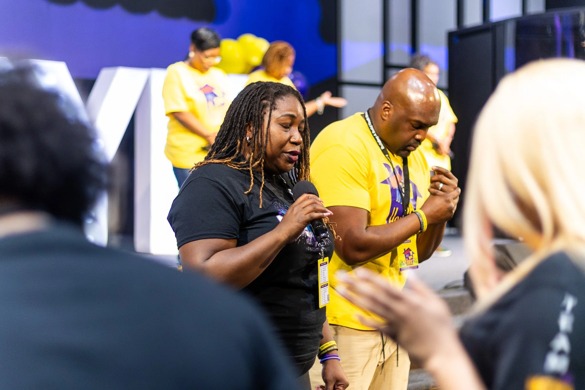 People praying with eyes closed, standing close together at an indoor event, some wearing yellow shirts with logos.