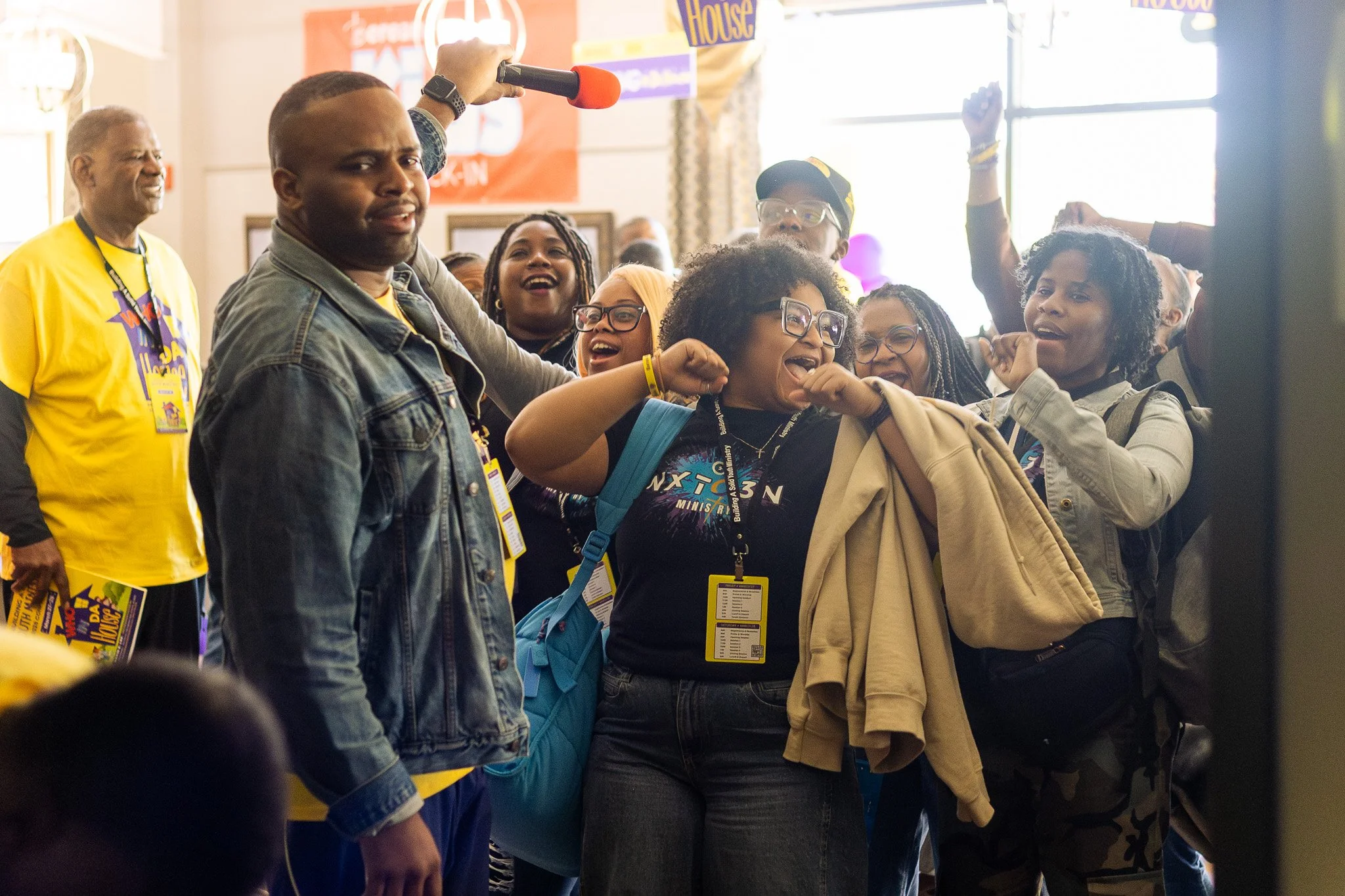 Group of people at an indoor event, some wearing yellow event badges, engaged in a lively activity or performance, with a man holding a microphone and others raising their fists or hands in the air.