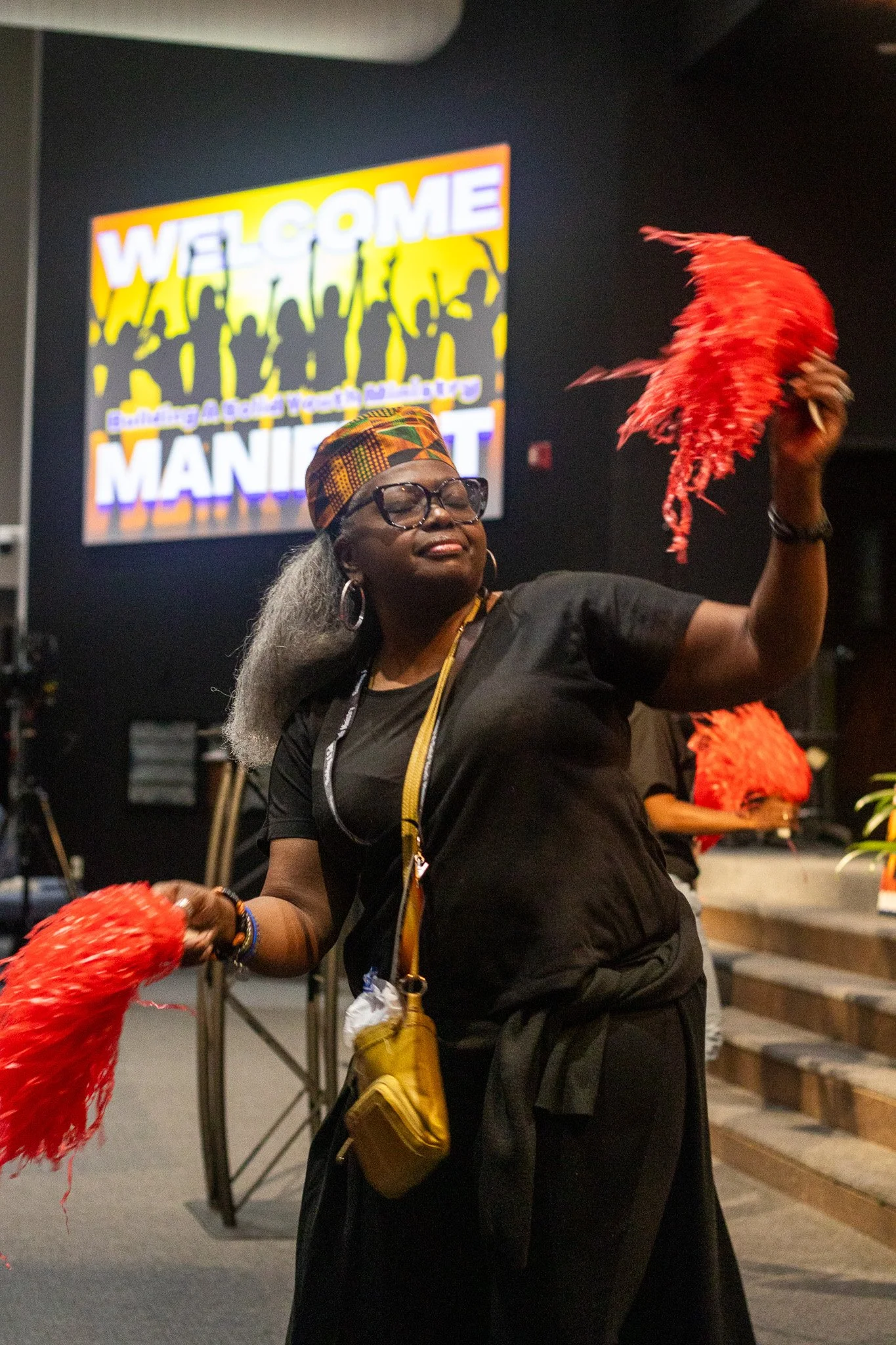 A woman with gray hair, glasses, and dark skin wearing a colorful headscarf, earrings, and a black outfit, holding a red feathered fan in her right hand, standing in front of a large sign with the word 'WELCOME' and silhouettes of people celebrating.