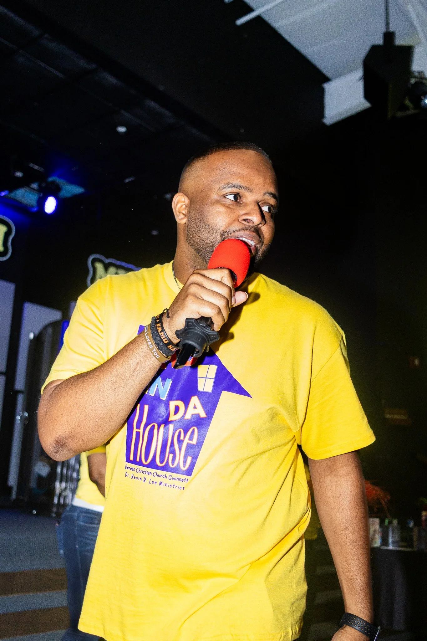 A man with a short haircut and beard holding a red microphone, wearing a yellow T-shirt with a graphic, at an indoor event with dark background and stage lighting.
