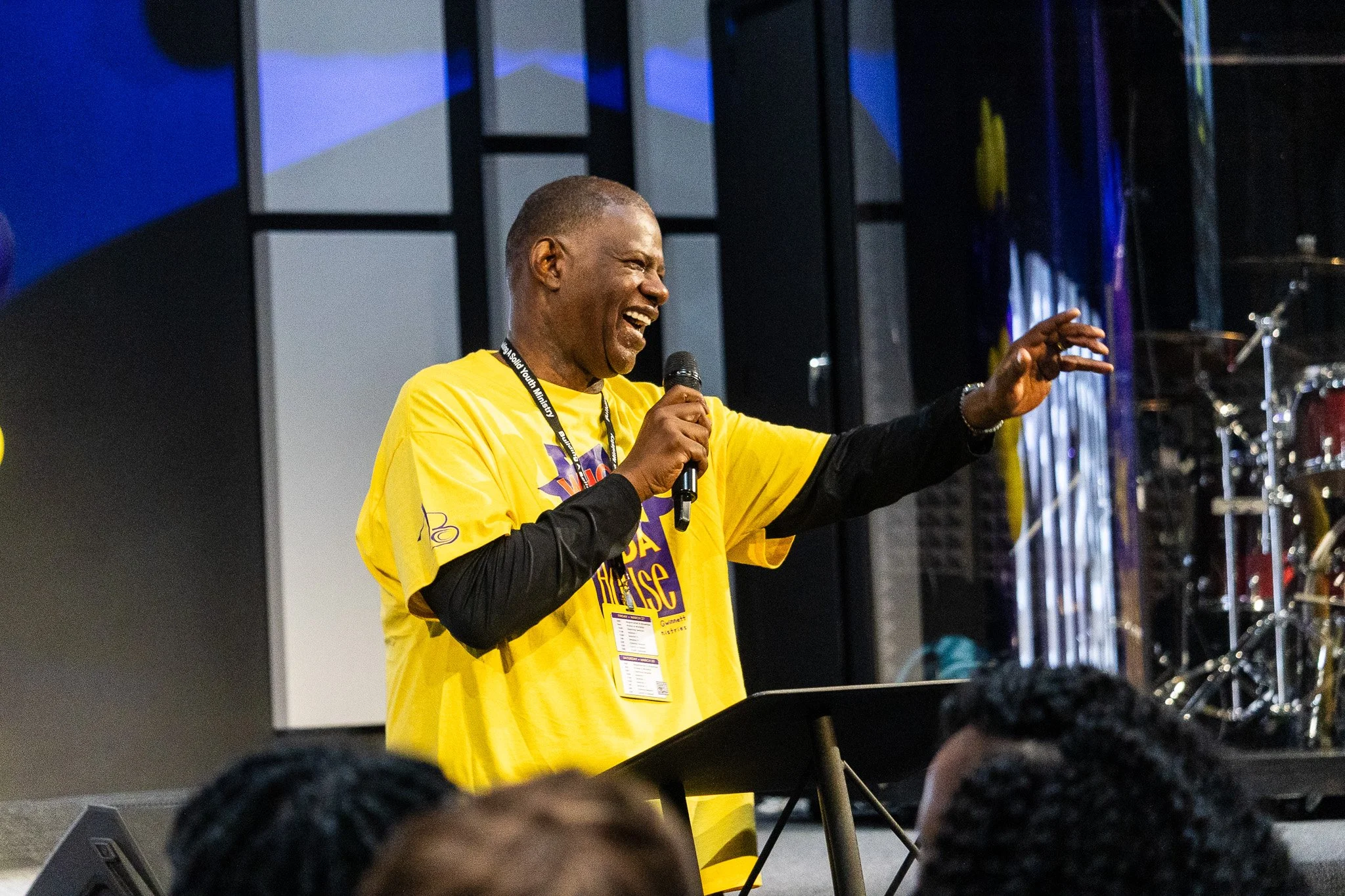A person wearing a yellow shirt and a black lanyard is speaking or singing into a microphone on a stage, with a bright smile and an expressive gesture. There are musical instruments and stage equipment in the background.