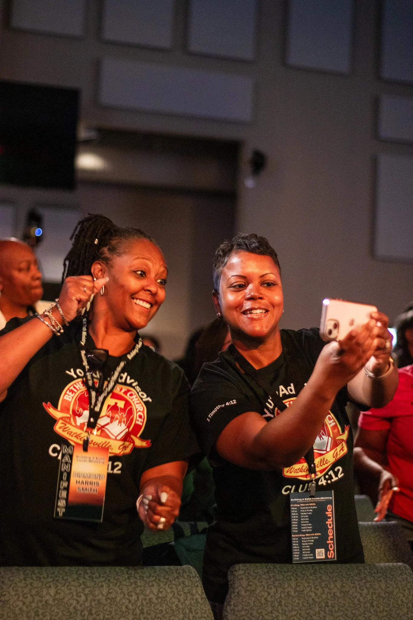 Two women are smiling and taking a selfie at an event. They are wearing black event T-shirts with a logo that says 'Watkinsville, GA' and some text. They have name tags and are standing in front of a dark background with a group of people.