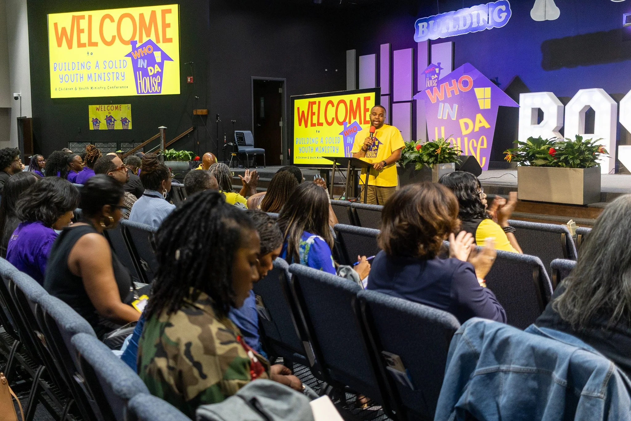 A man giving a speech at a youth ministry conference with a microphone, standing at a podium. The background includes colorful signs and decorations with the words 'WELCOME,' 'BUILDING A SOLID YOUTH MINISTRY,' and 'WHO IN DA HOUSE.' The audience cons