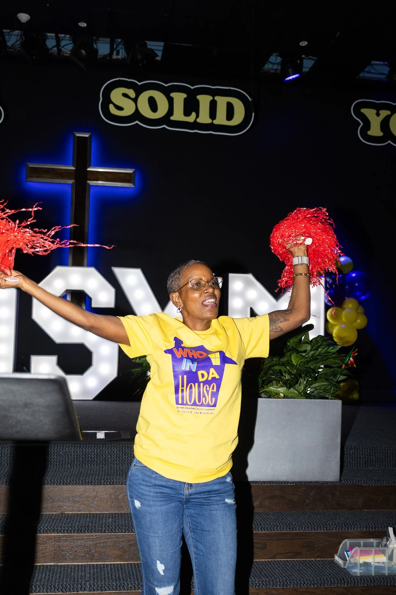 A woman wearing glasses and a yellow shirt with a logo that reads 'Who In Da House' is raising her arms and holding red pom-poms in a church or event setting. There are decorative balloons and a cross in the background.