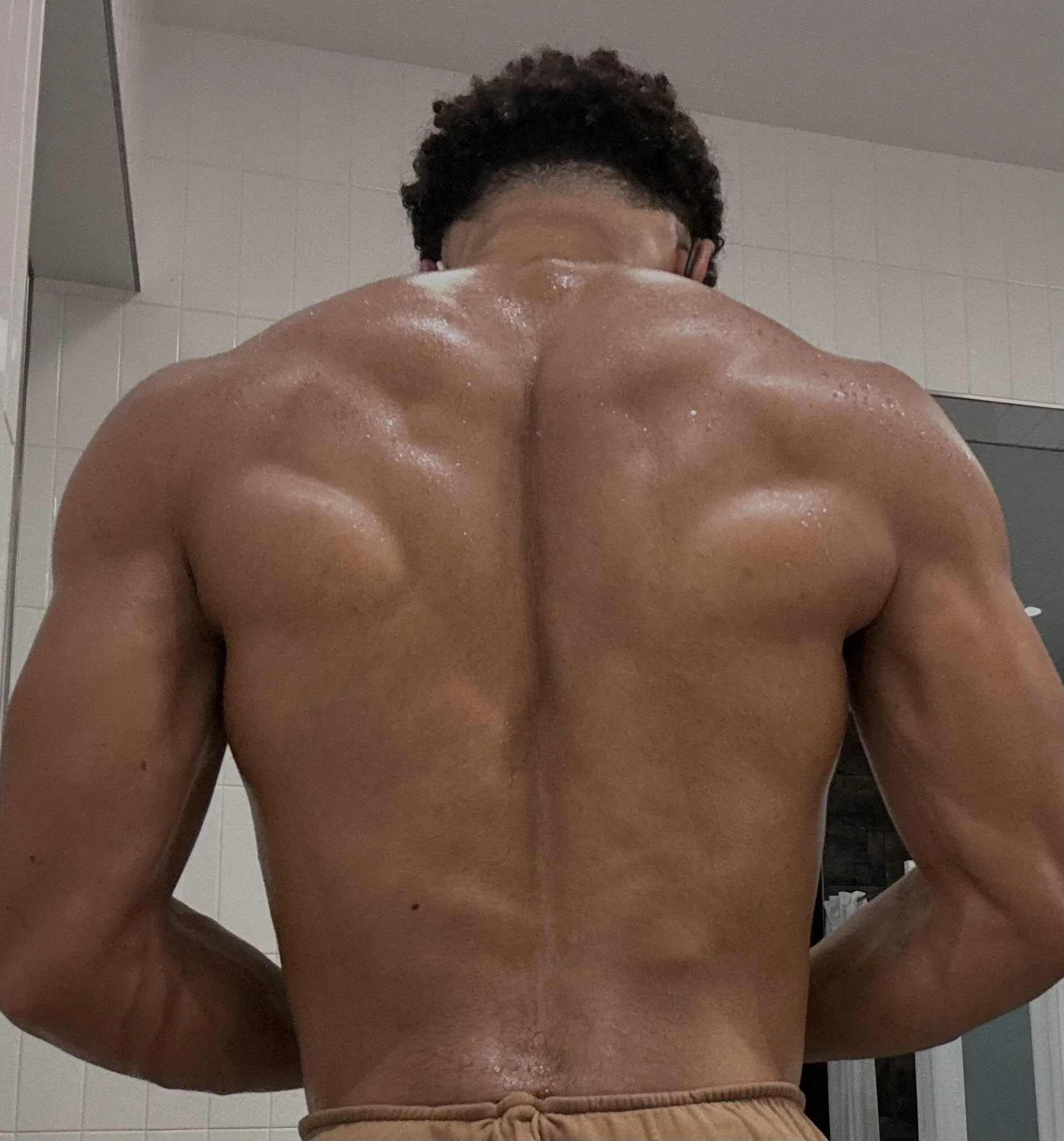 Back view of a muscular man with wet skin showing defined shoulder and back muscles, wearing beige shorts in a tiled room.
