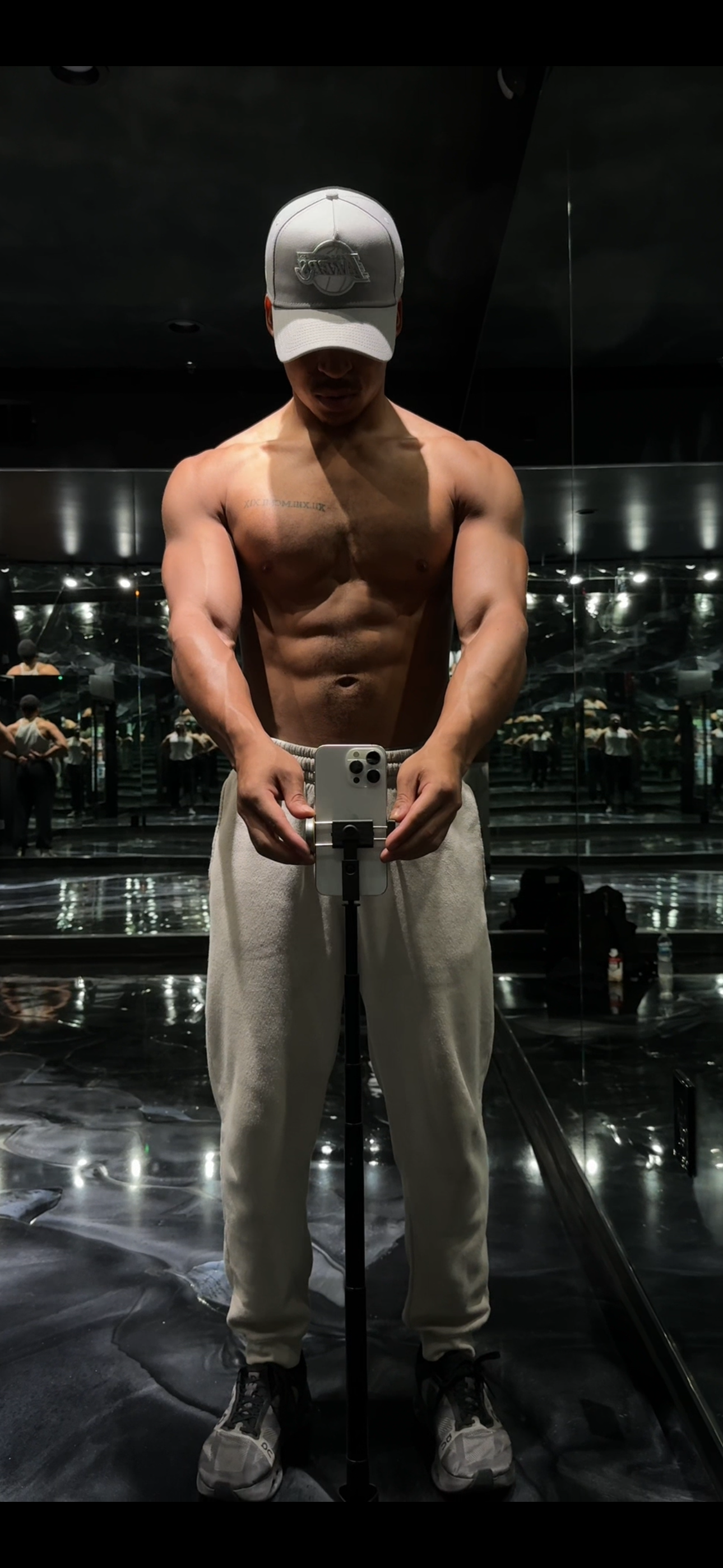 A shirtless man wearing a light gray cap takes a mirror selfie in a room with black walls and reflective surfaces. He is wearing beige sweatpants and gray sneakers, and his muscular build and tattoos are visible.