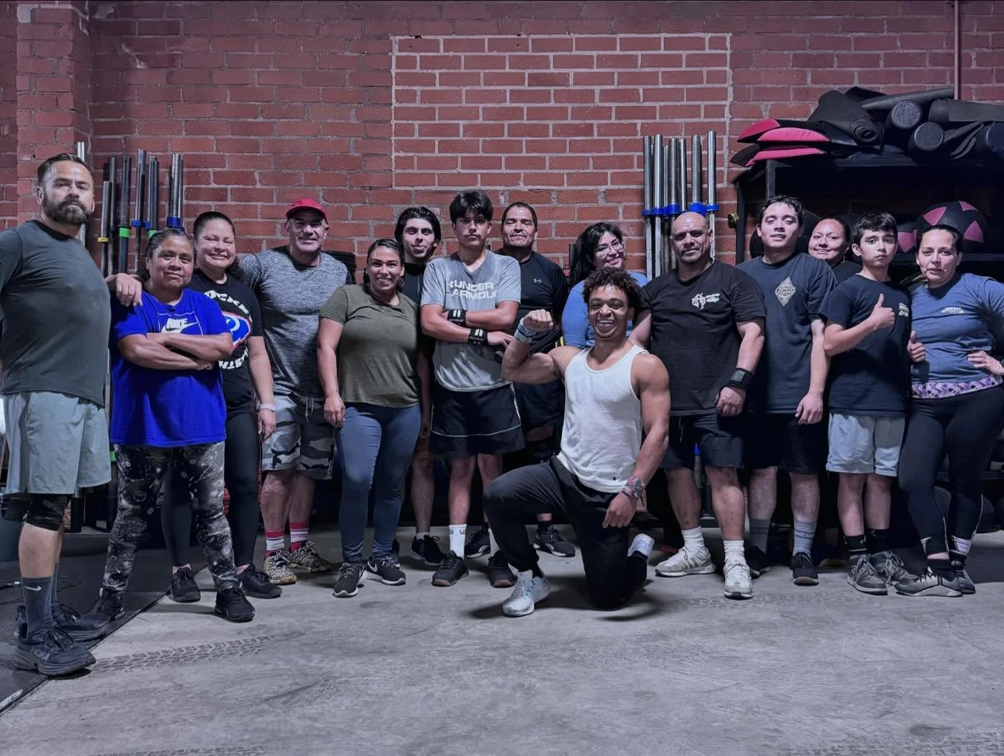 Group of people in workout gym, standing in front of brick wall, some flexing and others smiling, with fitness equipment like barbells and mats visible in background.