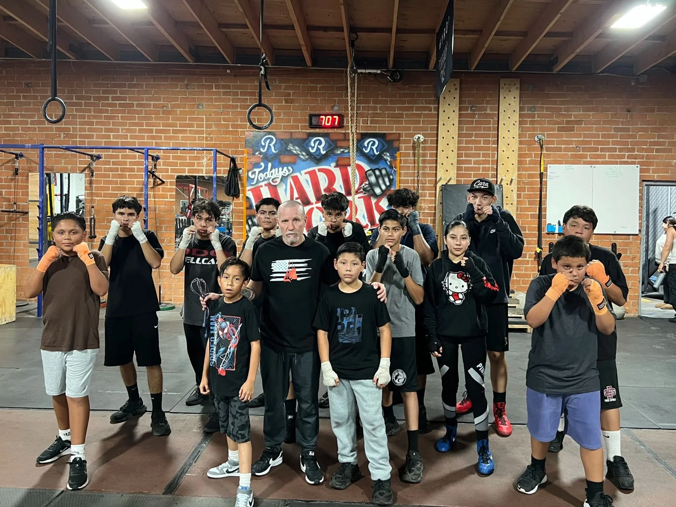 Boxing training session with a group of young boys and their coach in a gym. The boys are in fighting stance with gloves on, and the coach stands at the center. Gym equipment and a motivational sign are visible in the background.