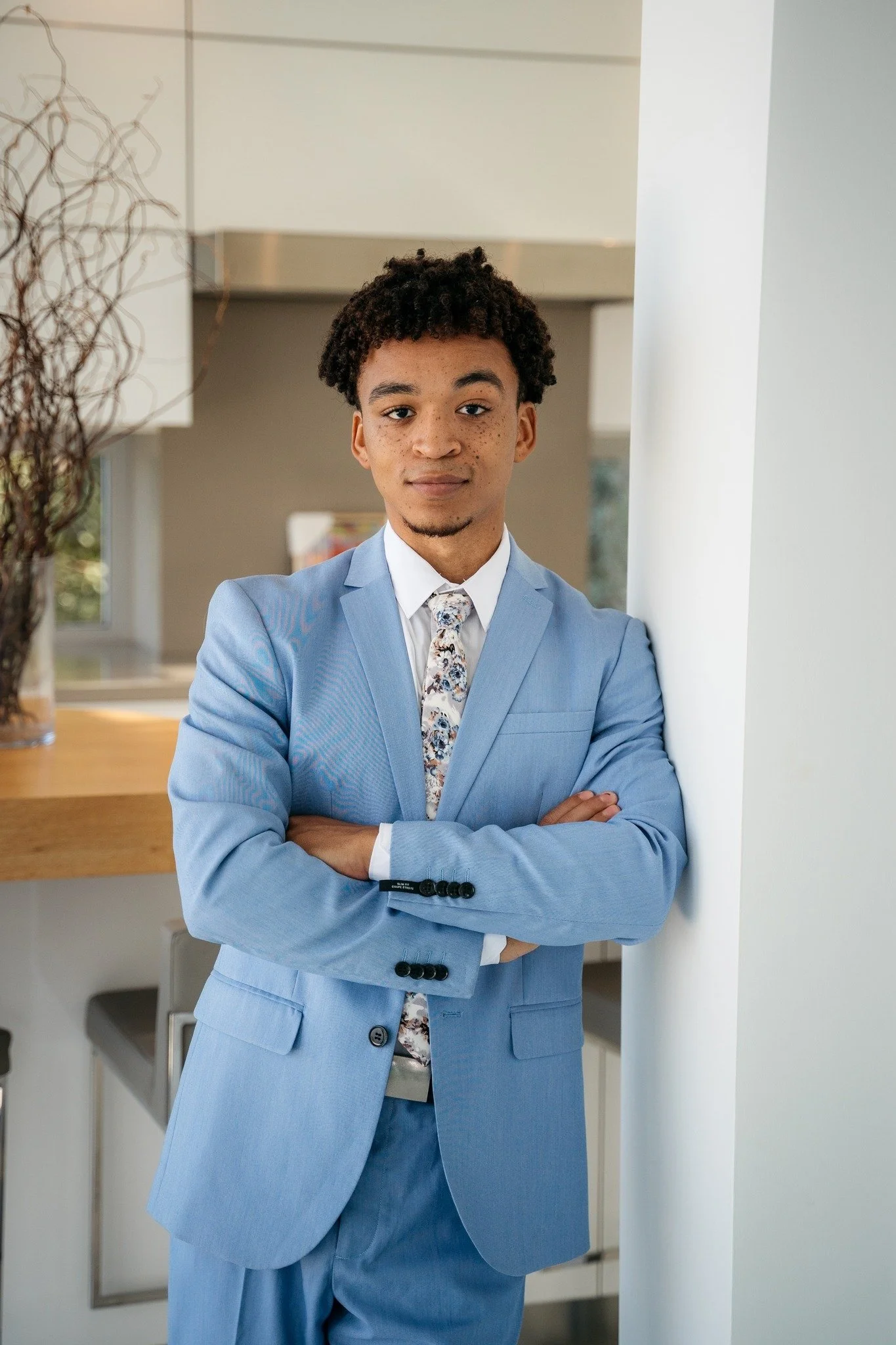 A young man with curly hair and freckles wearing a light blue suit, white shirt, and floral tie standing and leaning against a white wall with arms crossed in an indoor setting.