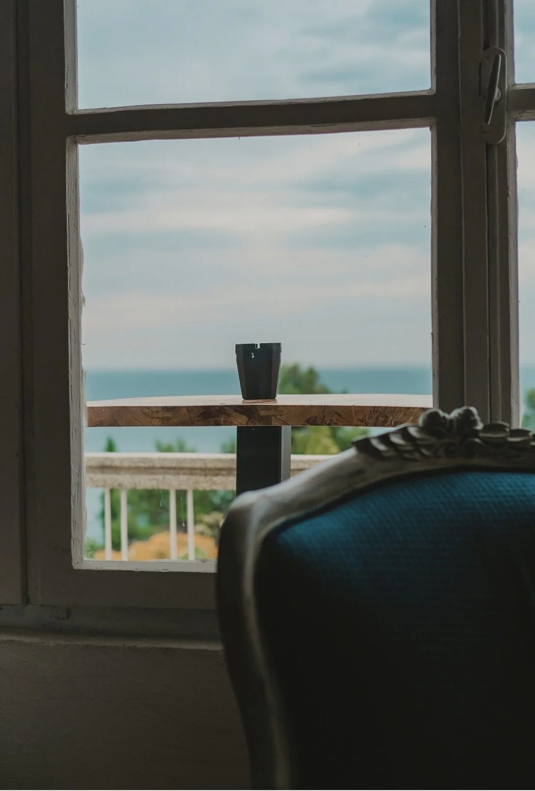 A view through a window showing a small table with a black cup on it, overlooking the ocean and sky in the background.