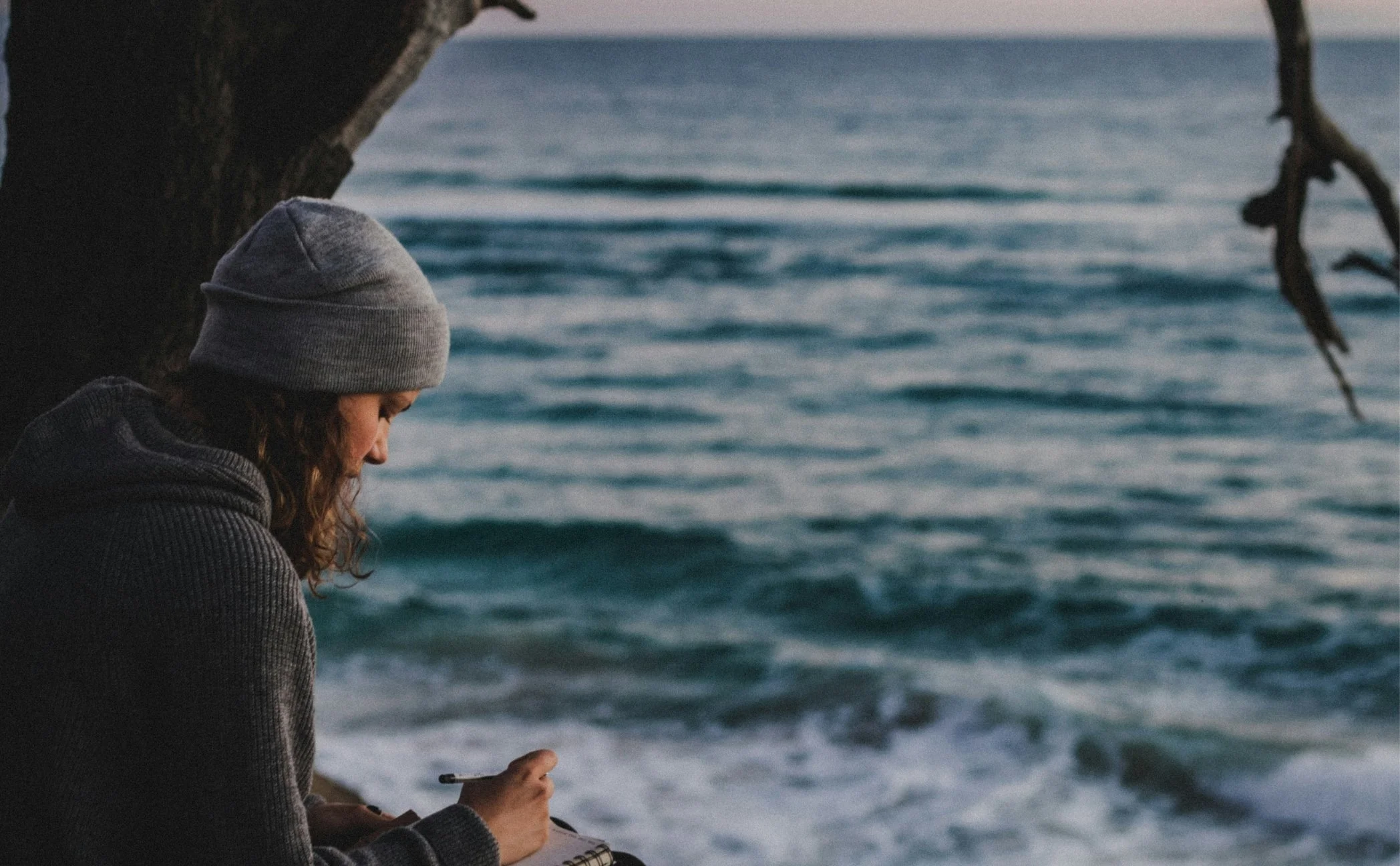 A young woman wearing a gray beanie and a dark gray hoodie sitting by the ocean, writing in a notebook with waves in the background.