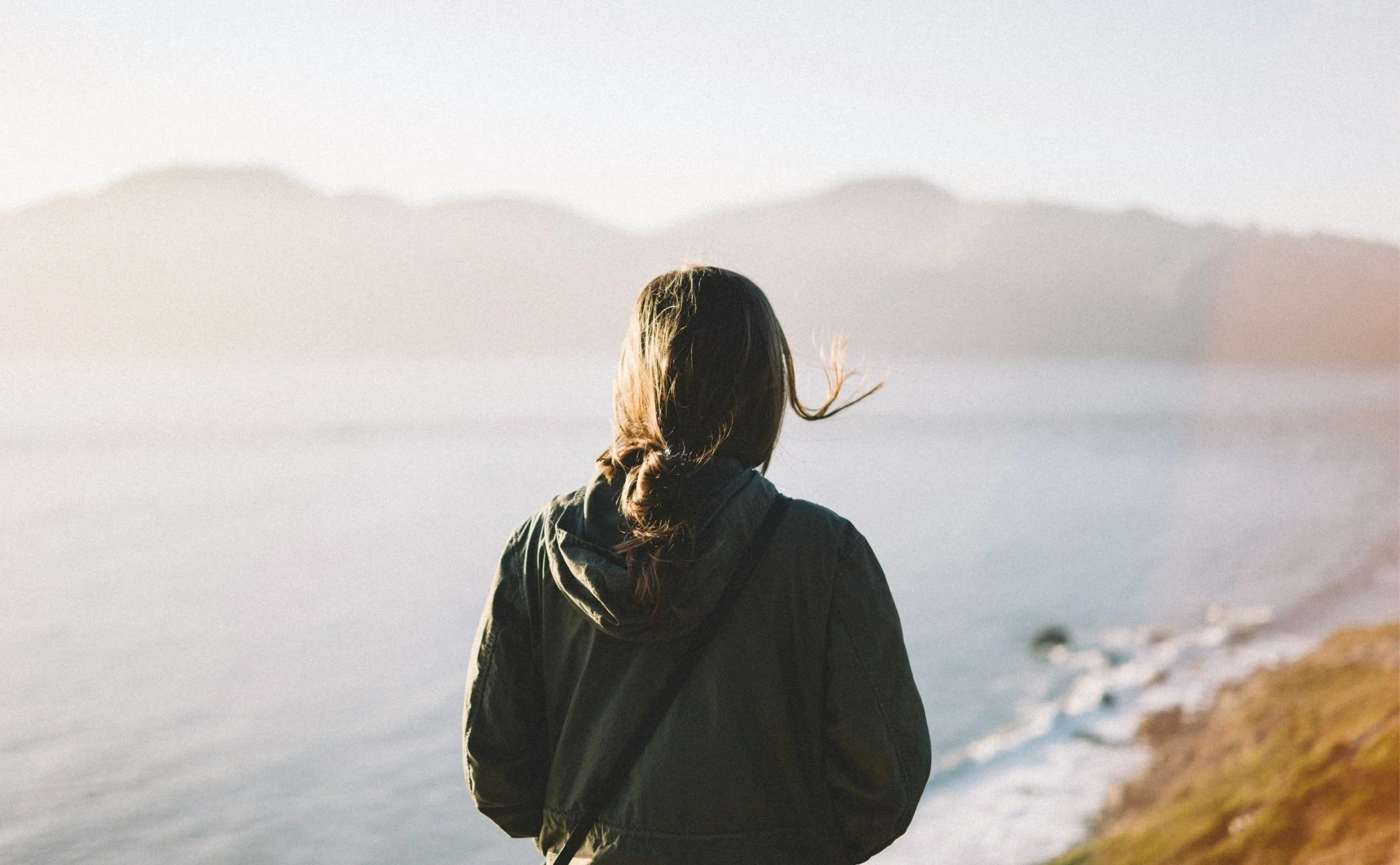 A woman with brown hair in a braid, wearing a green jacket, standing on a coastline during sunset, looking towards the ocean with distant mountains in the background.