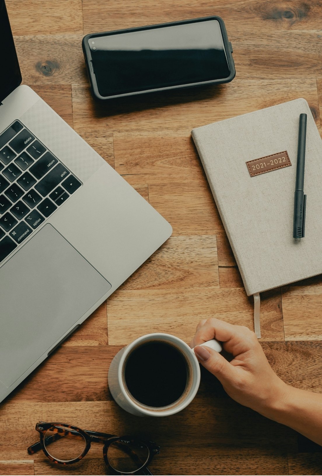 Desk with a MacBook, smartphone, planner for 2021-2022, black pen, cup of coffee, and tortoiseshell glasses.