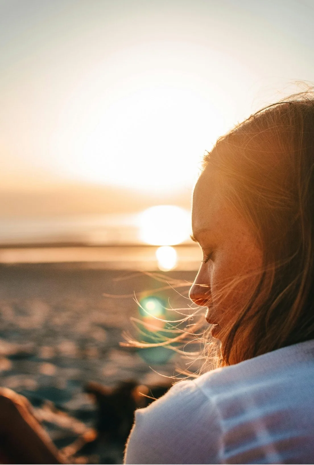 Side profile of a girl with long hair, sitting on the beach during sunset with her eyes closed.