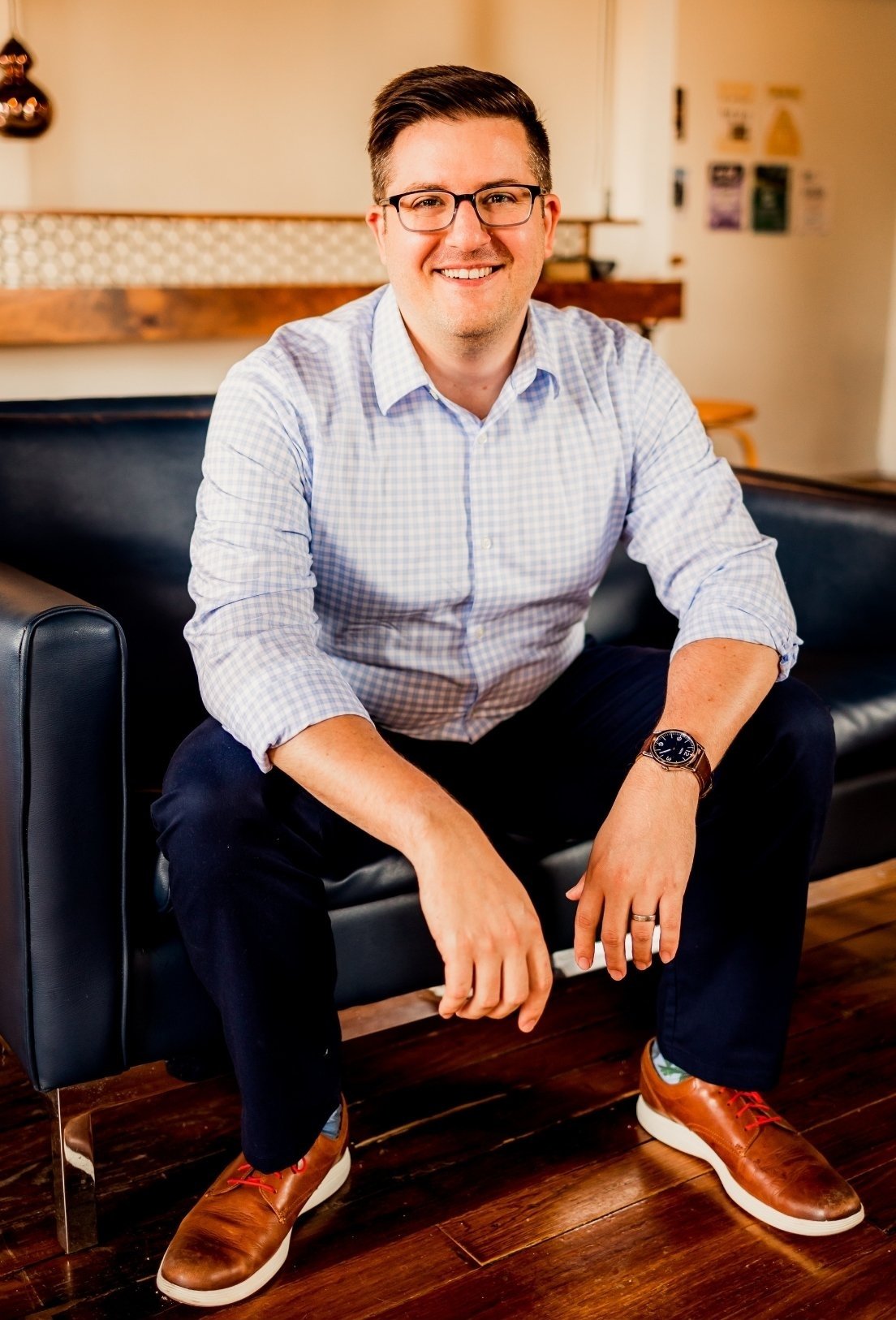 Joe Burke, Charlotte-based therapist, light blue checkered shirt, dark pants, brown shoes, and a watch, sitting on a black leather couch in a cozy room with wooden floors.