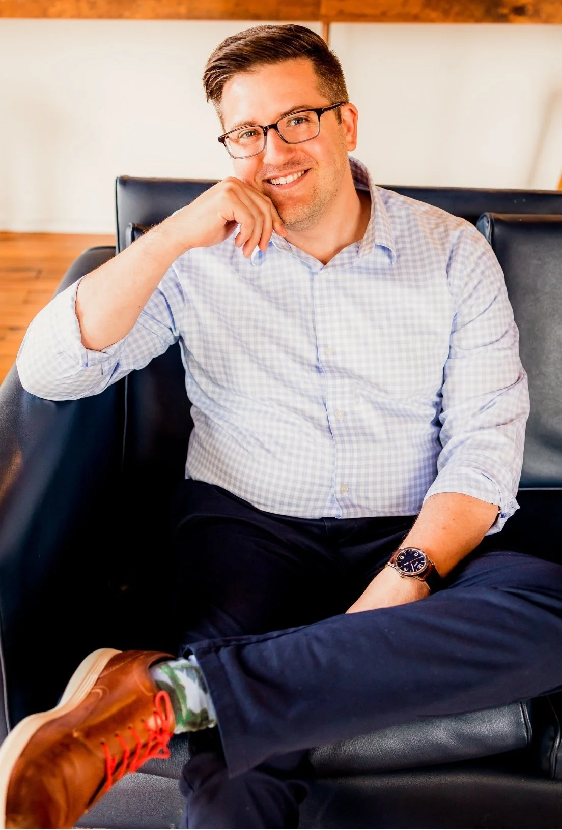 Charlotte therapist Joe Burke smiling, wearing glasses, a checkered shirt, a wristwatch, and brown shoes, sitting on a black leather couch.