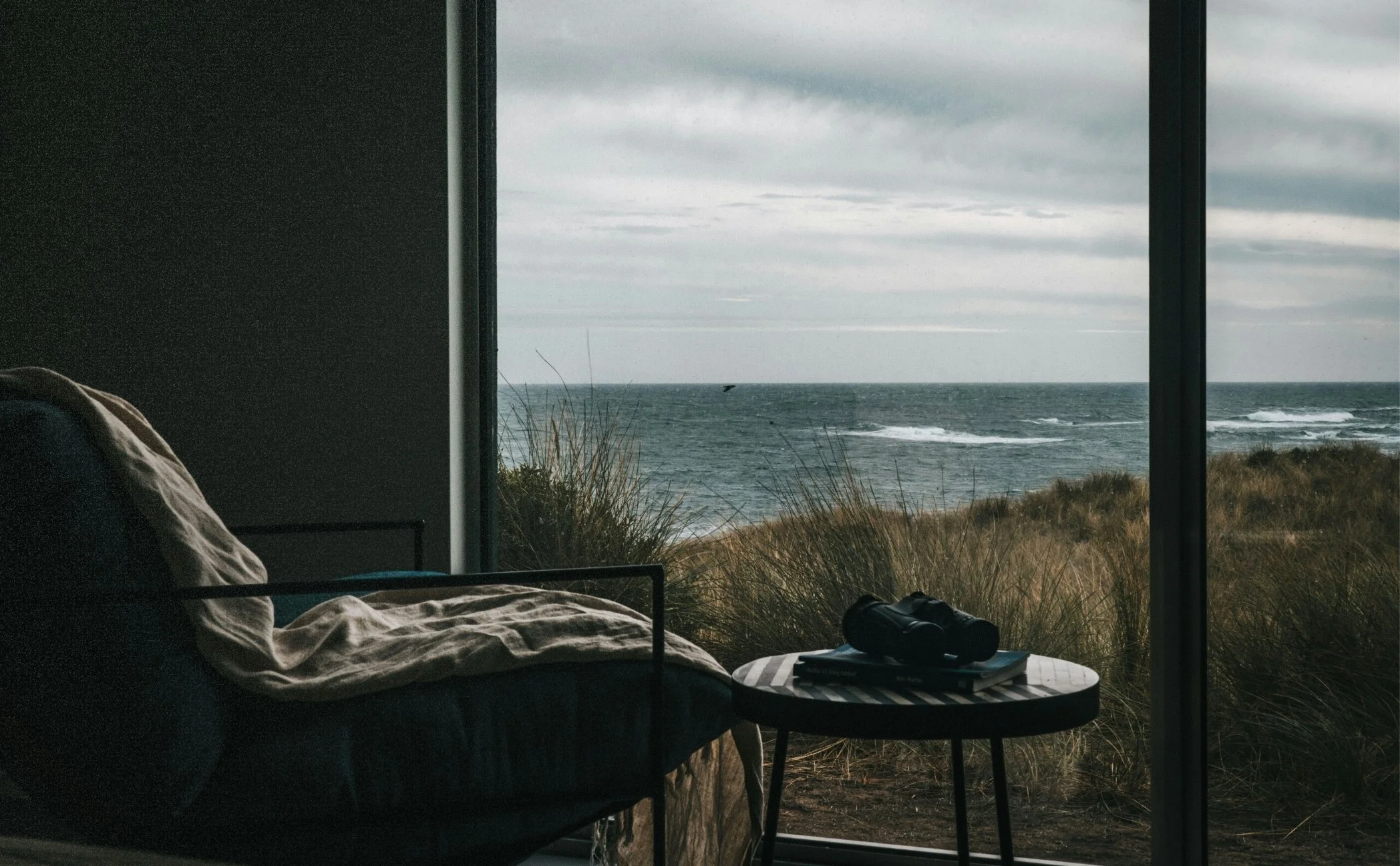 A cozy indoor space with a black armchair and a small round table, overlooking a beach through large glass windows. The table has binoculars and a magazine on it, and outside there are grassy dunes and the ocean under a cloudy sky.