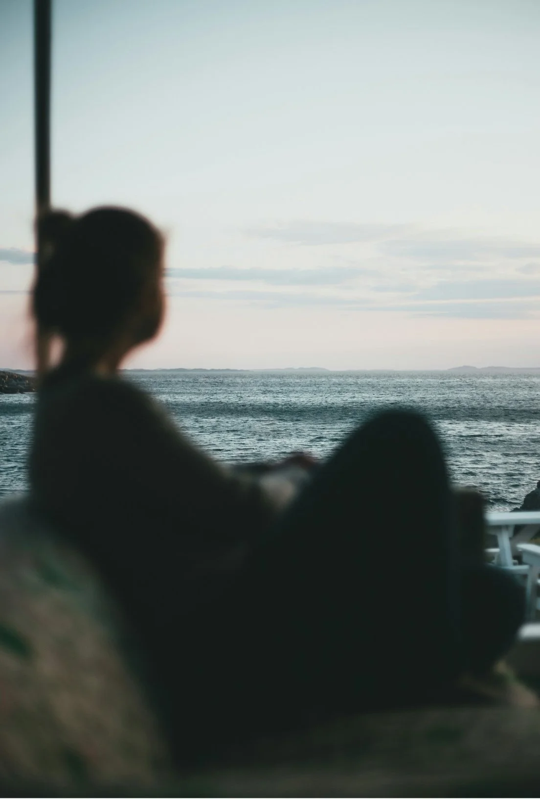 A person sitting indoors on a couch, looking out a large window at the ocean during sunset or sunrise.