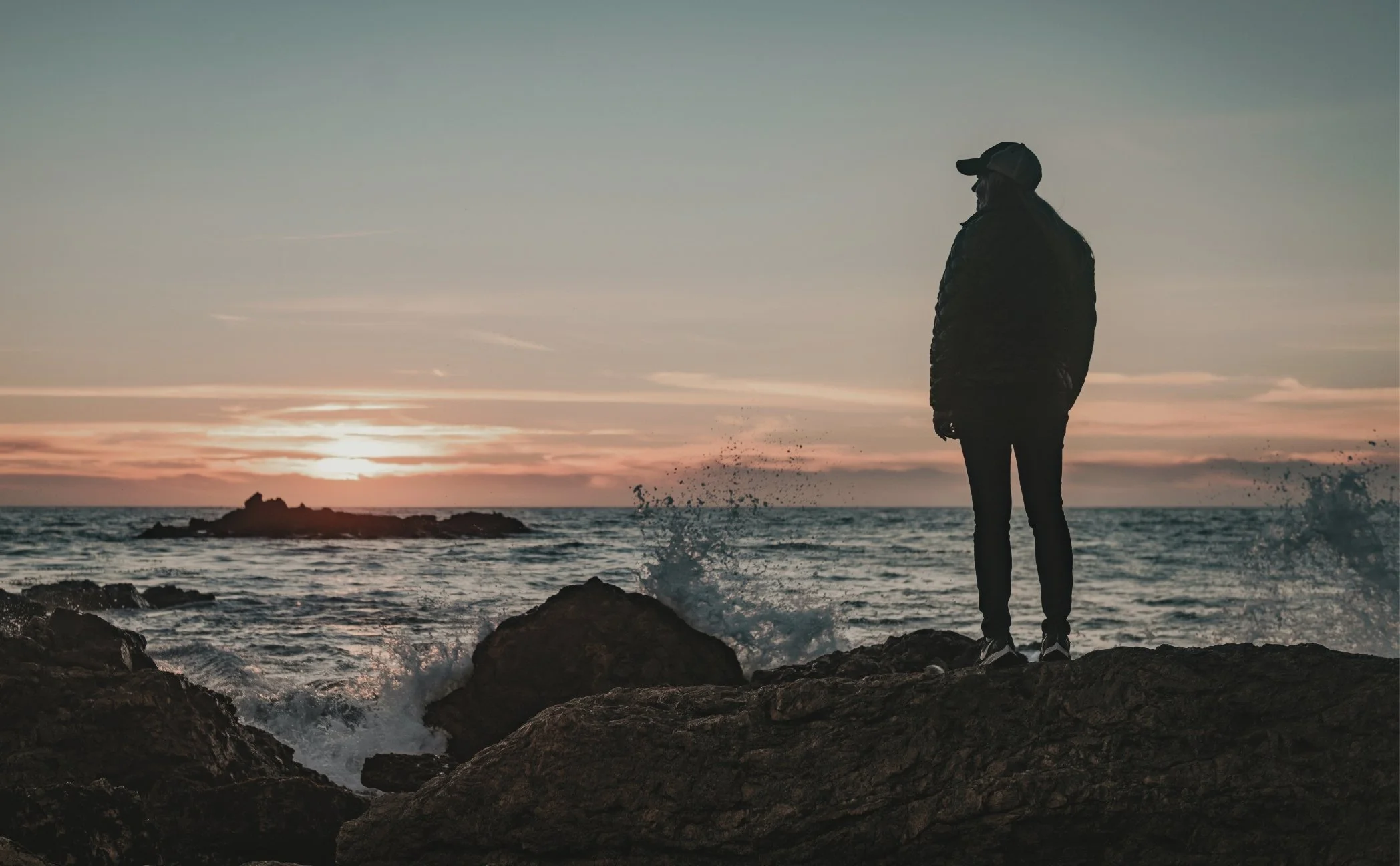 Person standing on rocky shoreline watching the sunset over the ocean