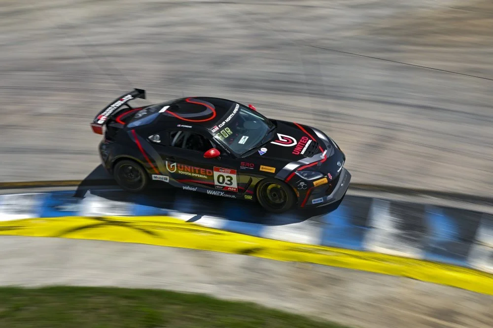 A black race car with red and yellow accents speeding on a race track, kicking up dust and veering over painted blue and yellow curb.