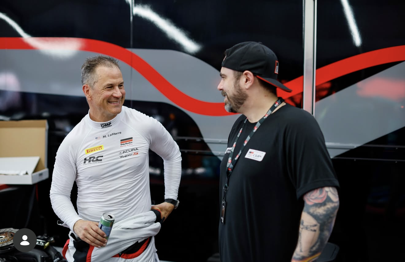Two men smiling and talking, one in a race car driver's suit holding a beverage, the other in casual black clothing with tattoos, standing near a black and red racing team trailer.